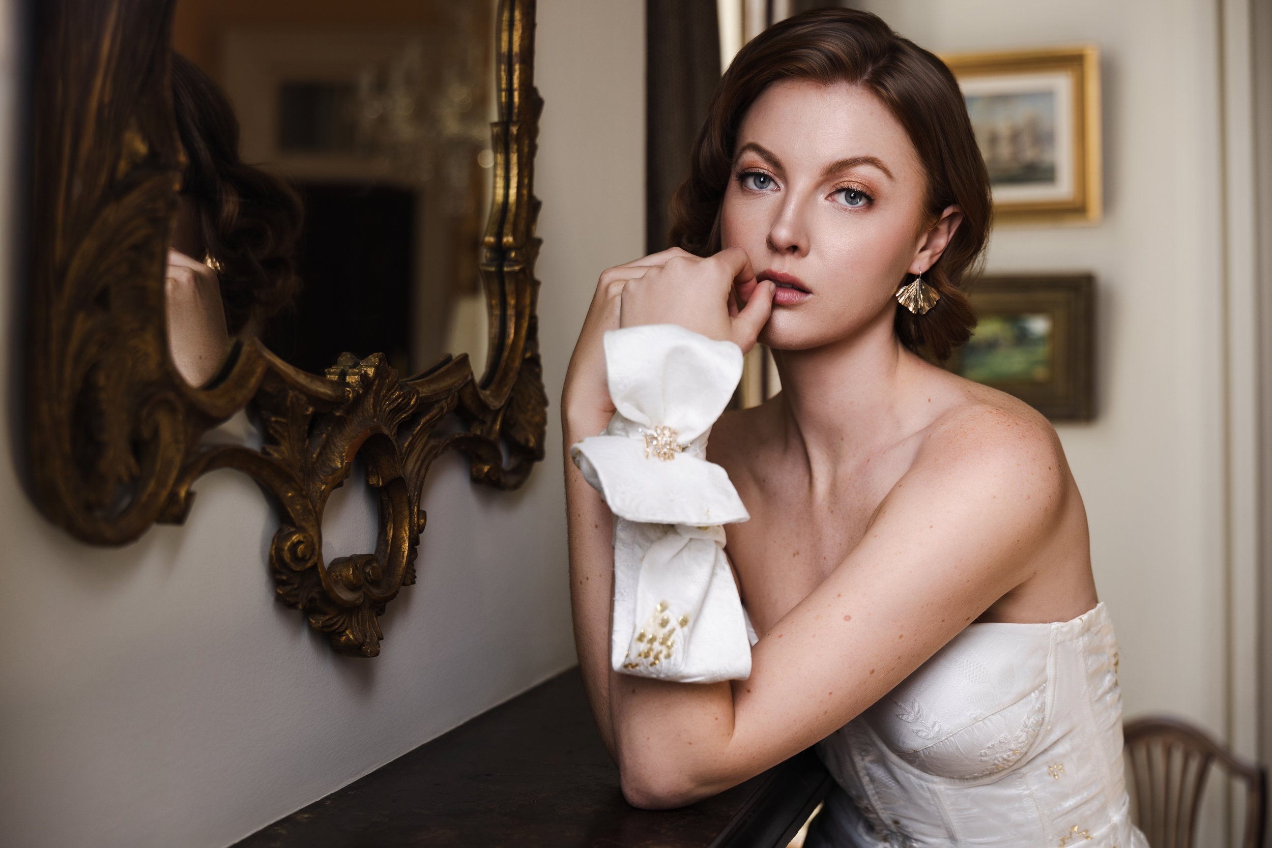 Bride poses with soft expression in vintage-inspired wedding look beside gilded mirror and fireplace