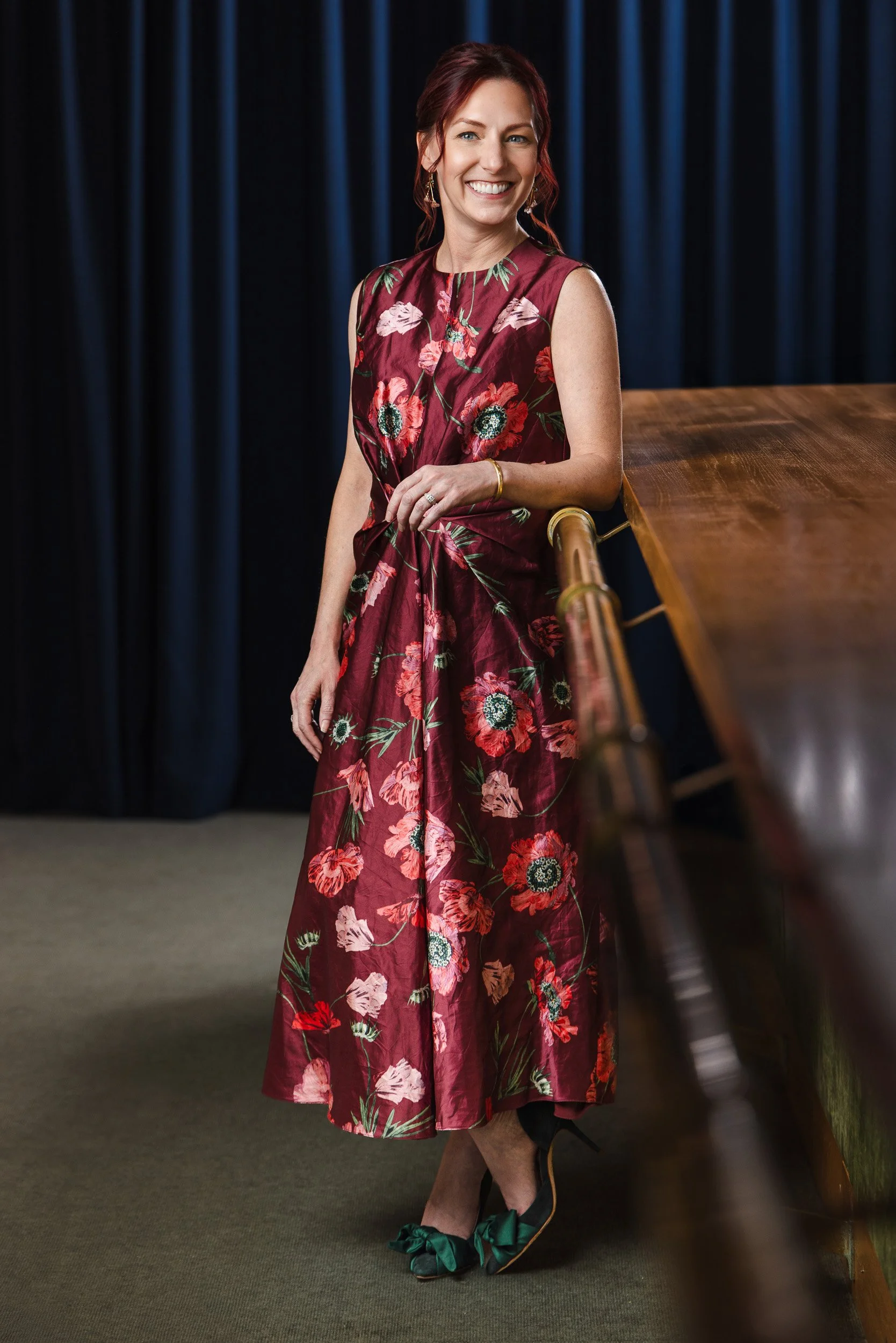 Carrie Kitko in a burgundy poppy print dress and green bow heels, leaning against a brass staircase railing for her Nashville editorial portrait.