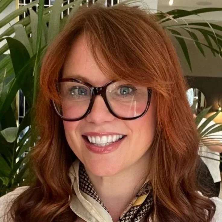 A woman with red hair, glasses, and a bright smile, standing in front of green plants.