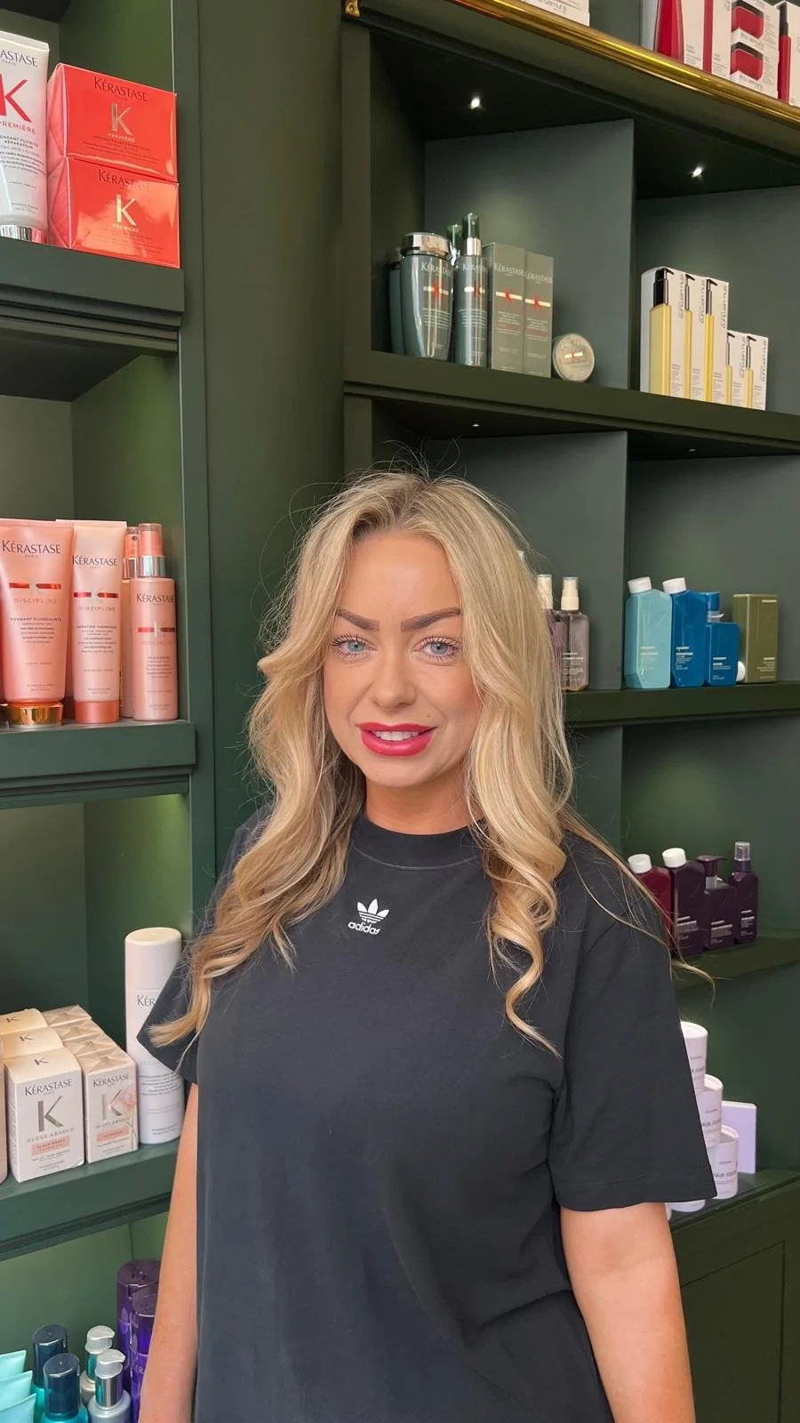 A woman with long blonde hair, blue eyes, and pink lipstick standing in front of green shelves stocked with hair and beauty products in a salon or store.