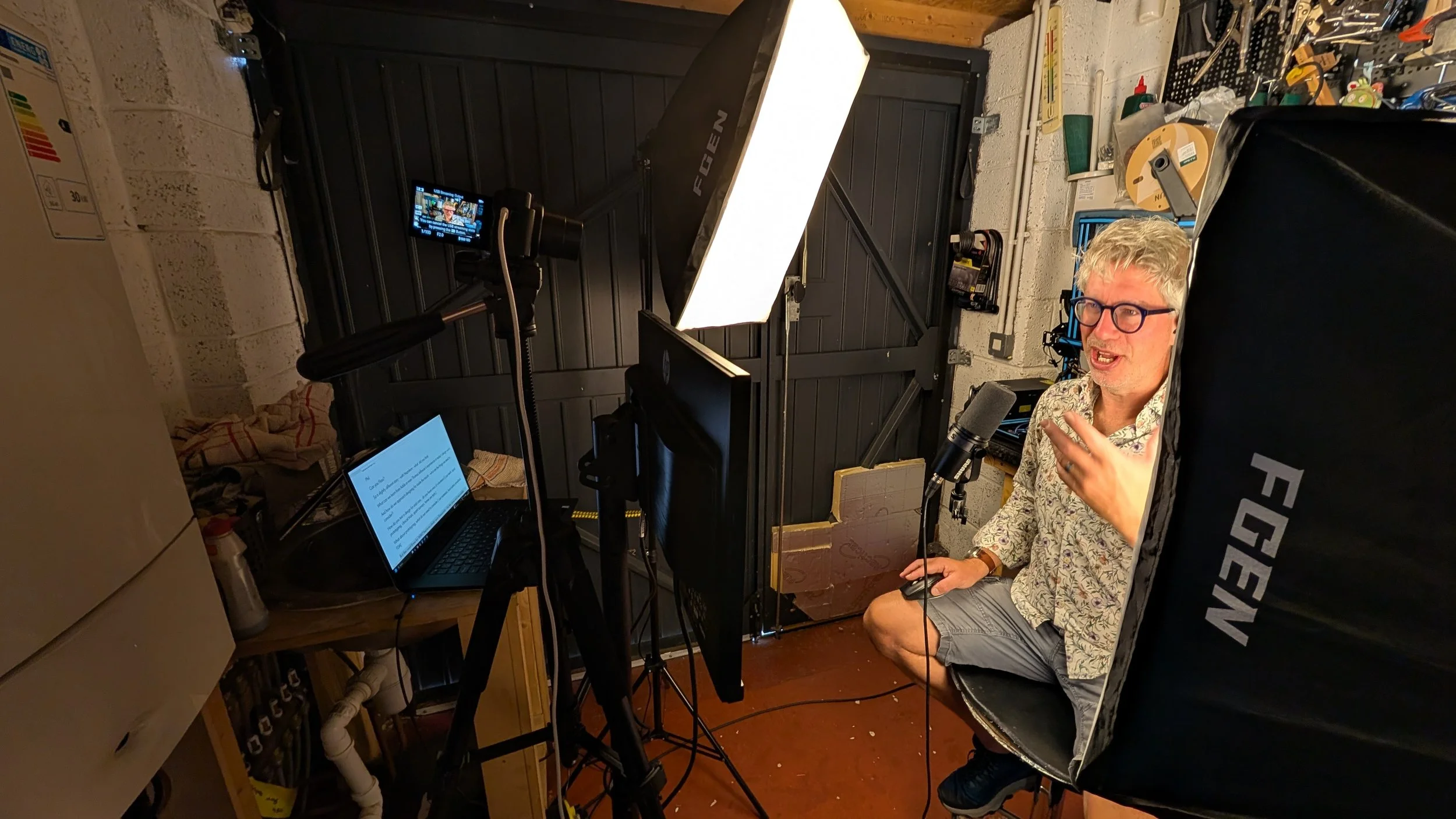 A man with gray hair, glasses, and a patterned shirt is sitting in a small recording studio or setup with a black backdrop. He is speaking into a microphone and gesturing with his hand. There is electronic equipment, a laptop, and a large softbox light in the scene.