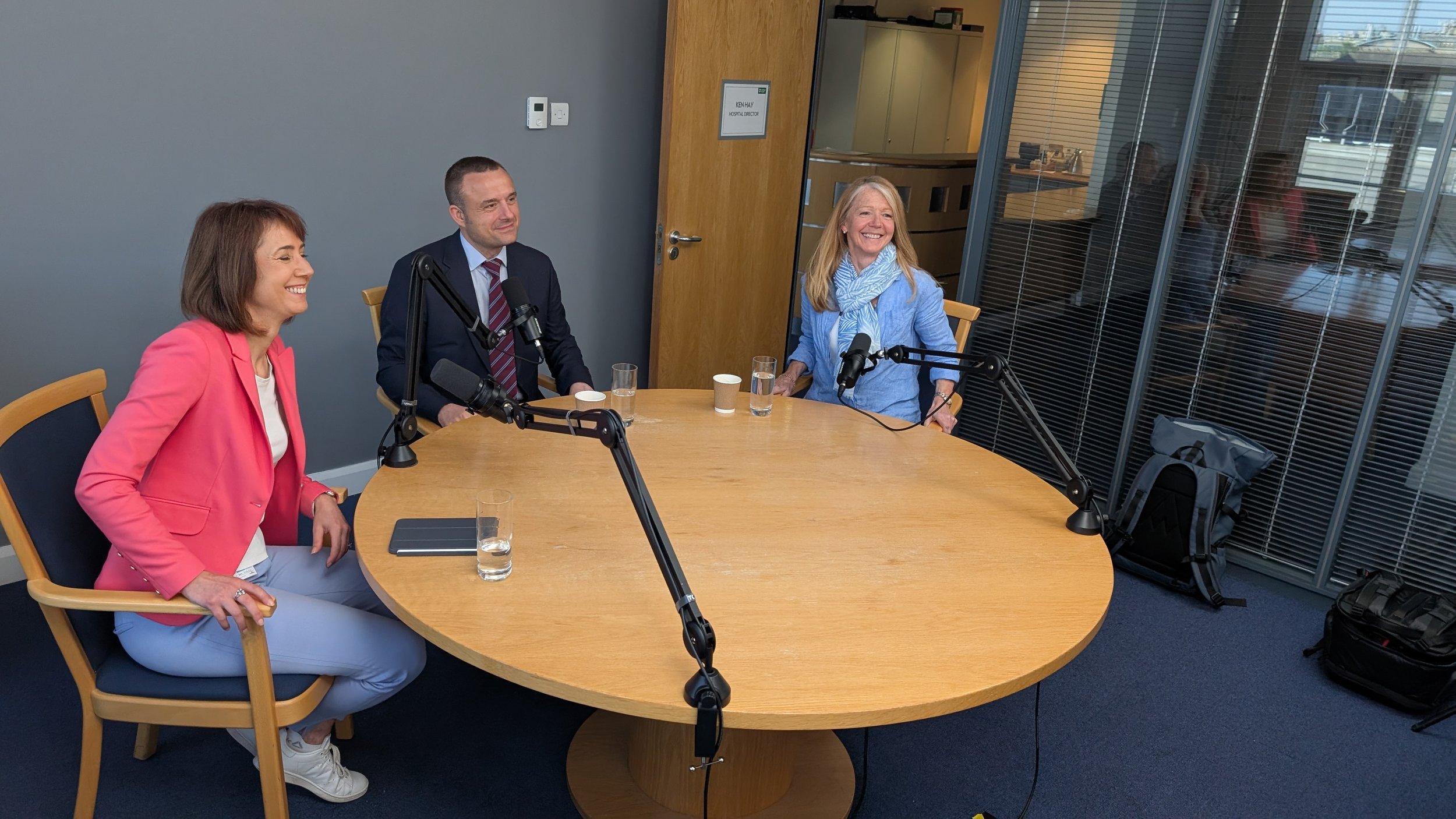 Three people sitting at a round table in a podcast recording studio, two women and one man, smiling and laughing, with microphones and glasses of water in front of them, in a modern office setting.