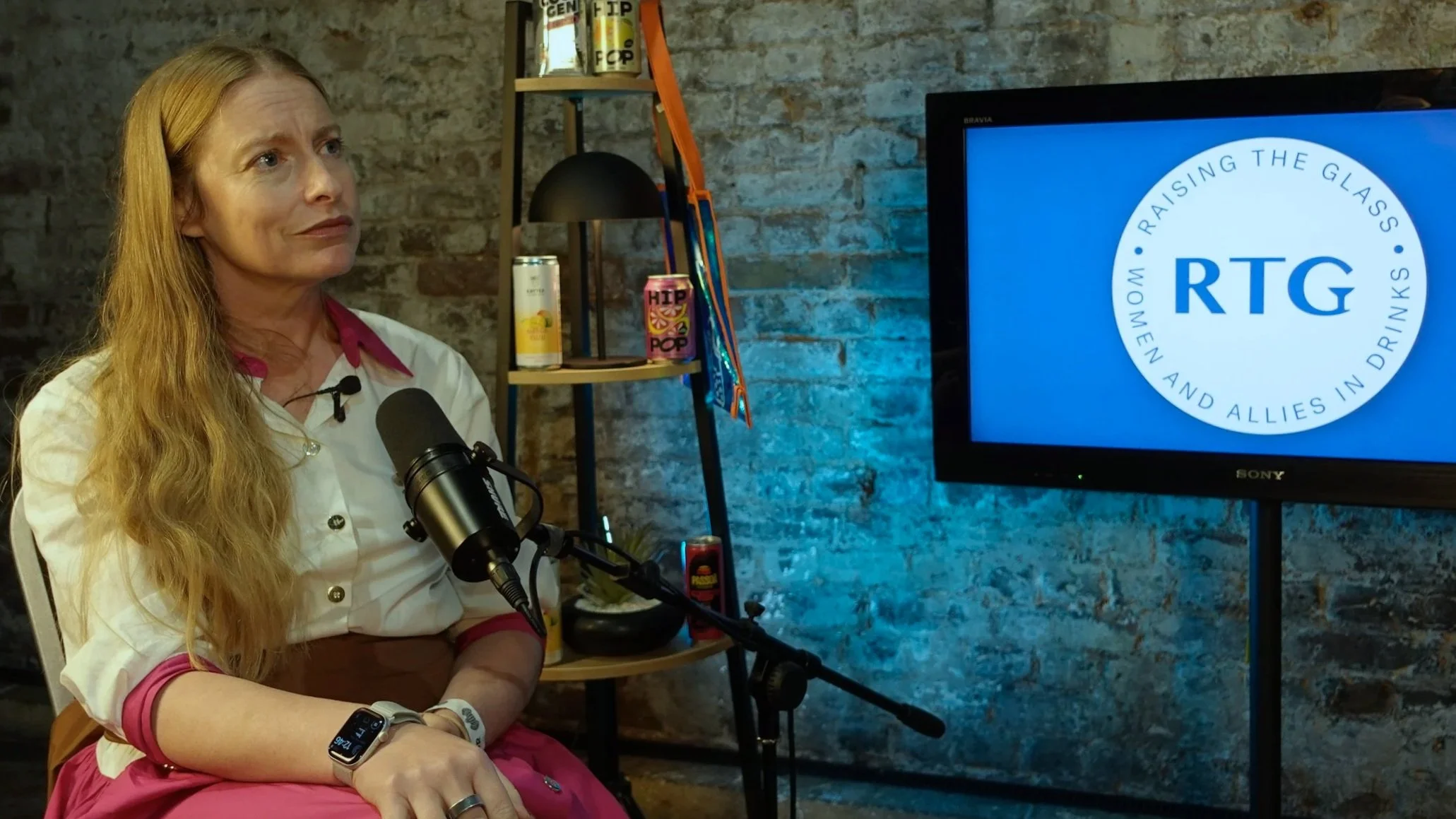A woman with long red hair sits in front of a microphone, in a room with a brick wall. A TV screen to her right displays a blue and white logo with the words 'Raising The Glass' and 'RTG' at the center.