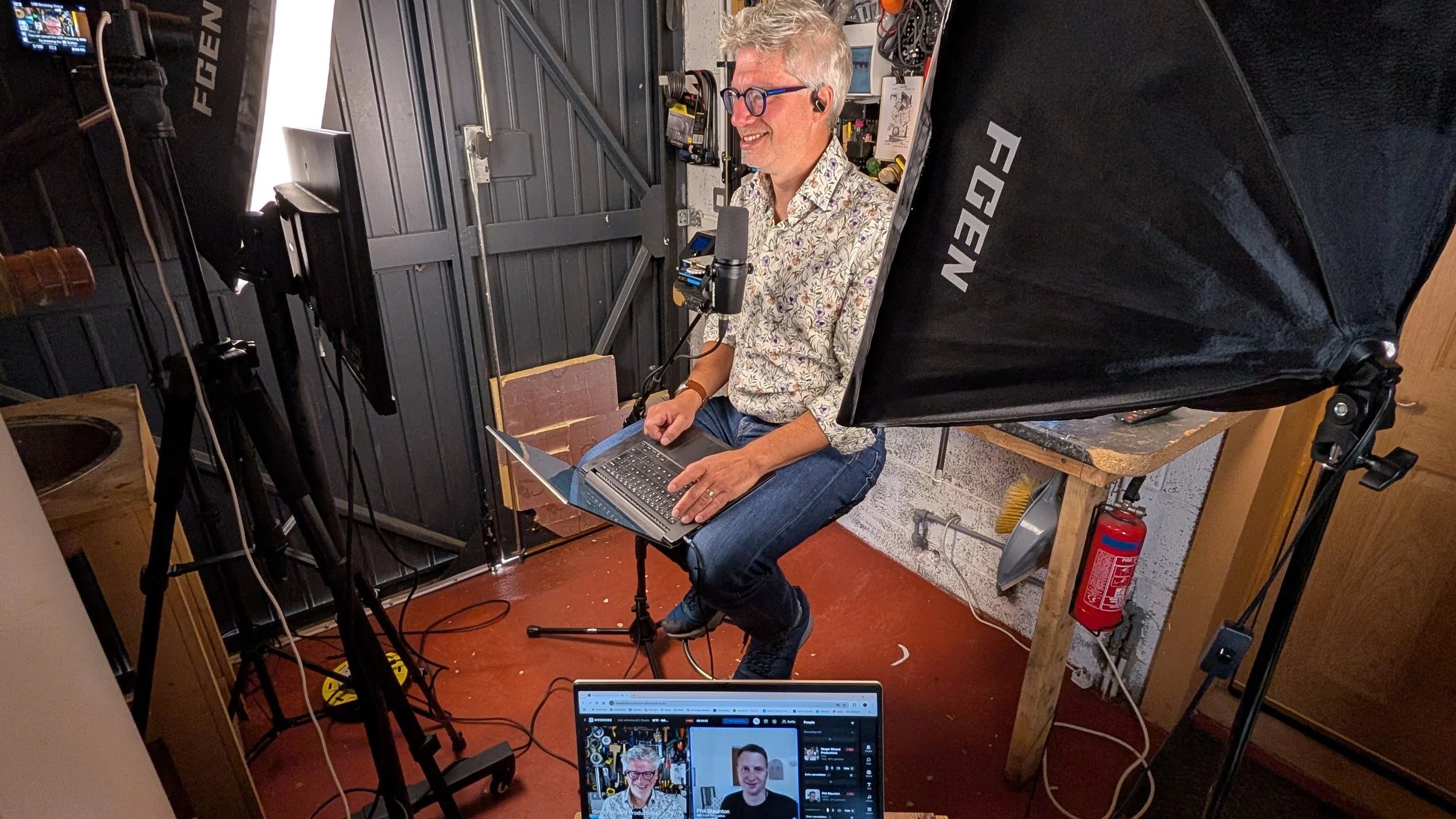 A man with glasses and gray hair, sitting on a chair in a studio, is smiling and operating a laptop. He is surrounded by professional camera and lighting equipment, recording or streaming a video or podcast, with a monitor showing his image.