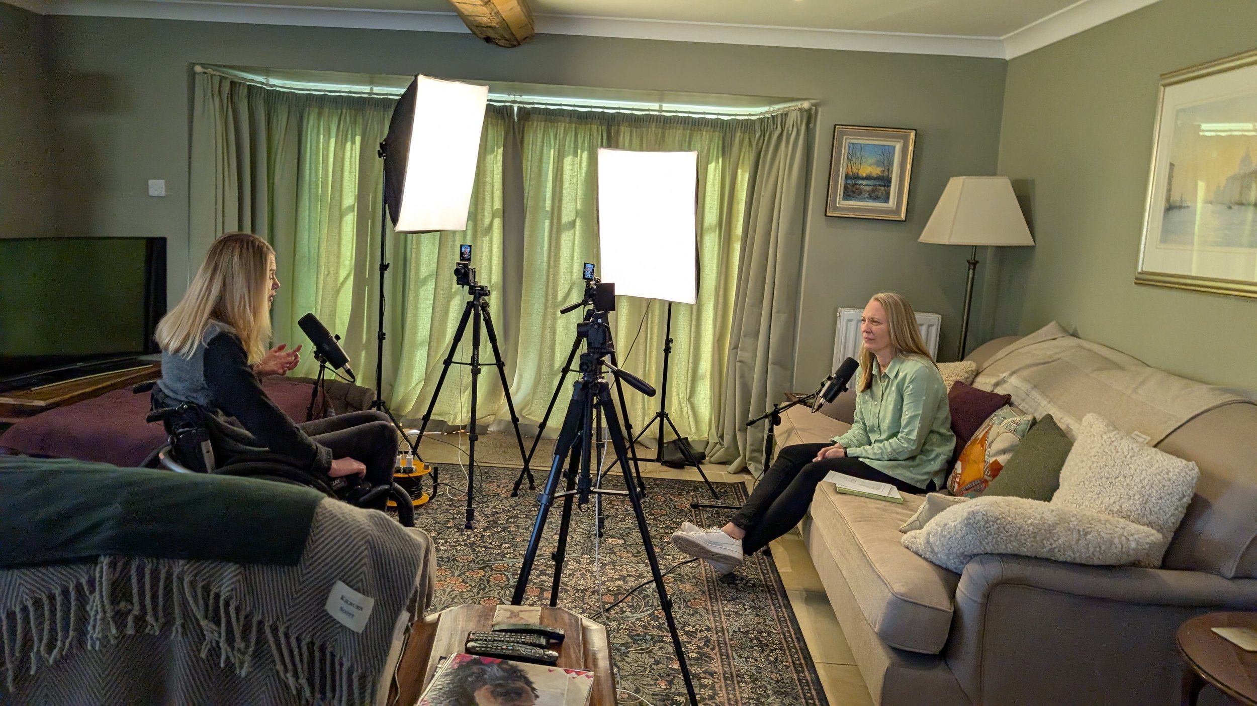 Two women sitting in a living room being interviewed or recording a podcast with cameras and lighting equipment set up between them.
