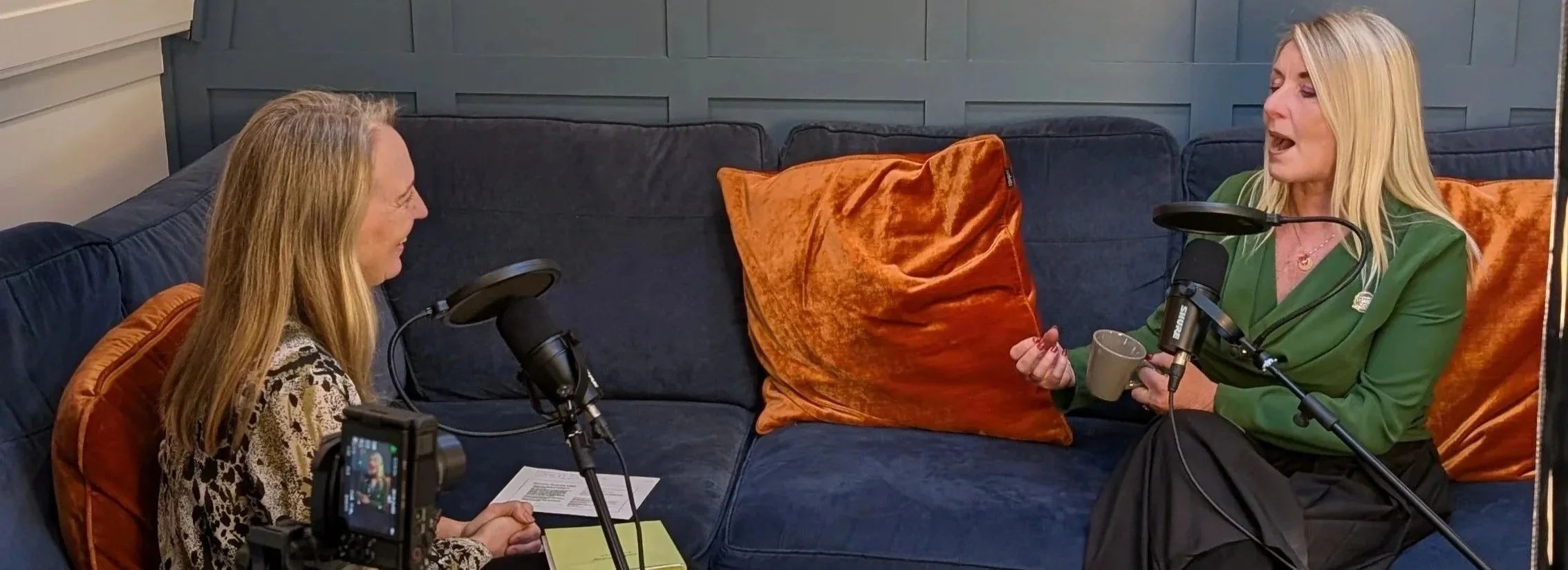 Two women with long blonde hair, sitting on a blue couch with orange pillows, having a podcast recording session. Both have microphones. One woman in leopard print top holds notes, and the other in green top with a mug is speaking.