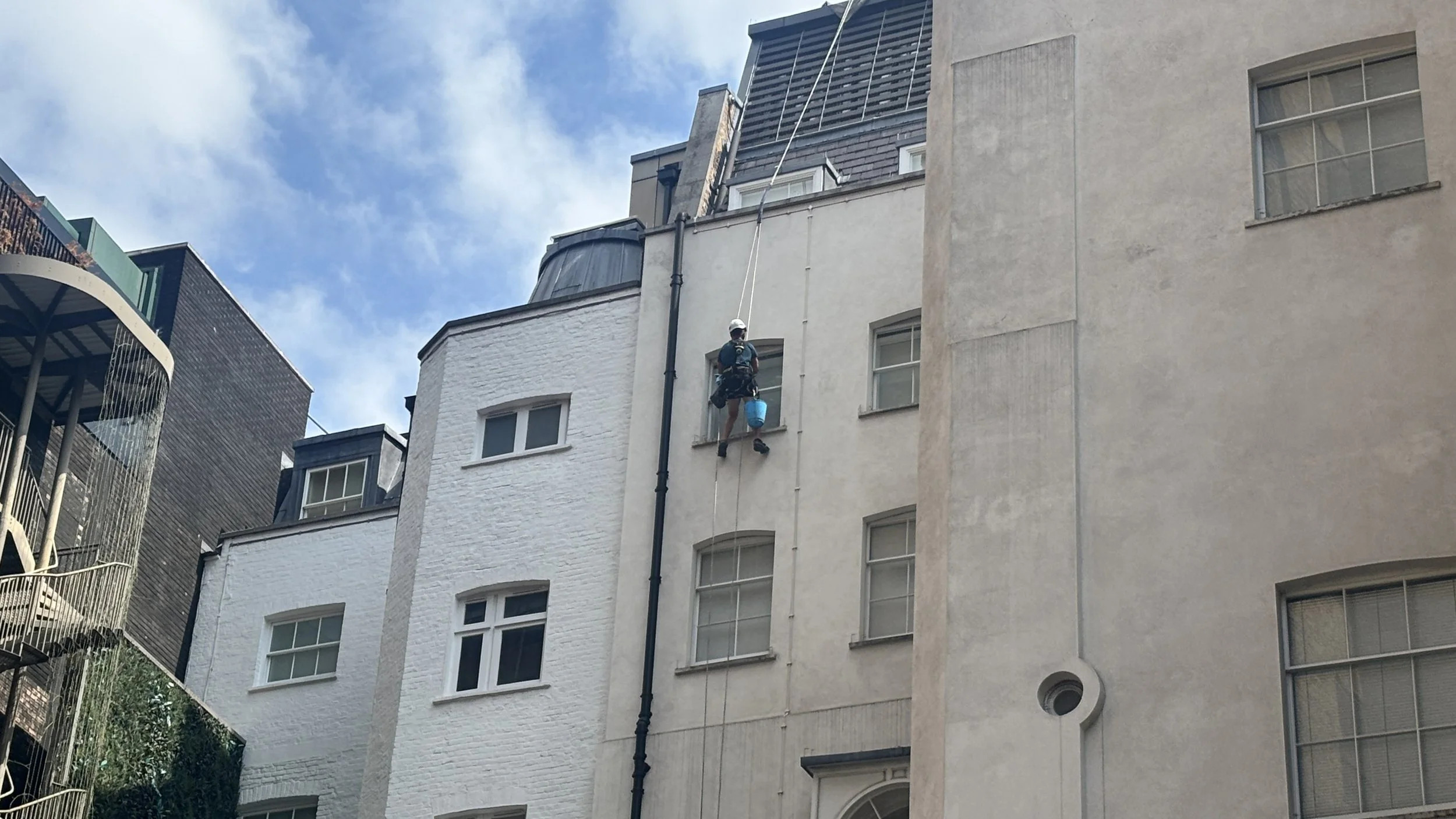 A person wearing a helmet and harness is climbing or working on the exterior of a multi-story building using a rope. The building has several windows, and other buildings with modern architecture are visible nearby under a partly cloudy sky.