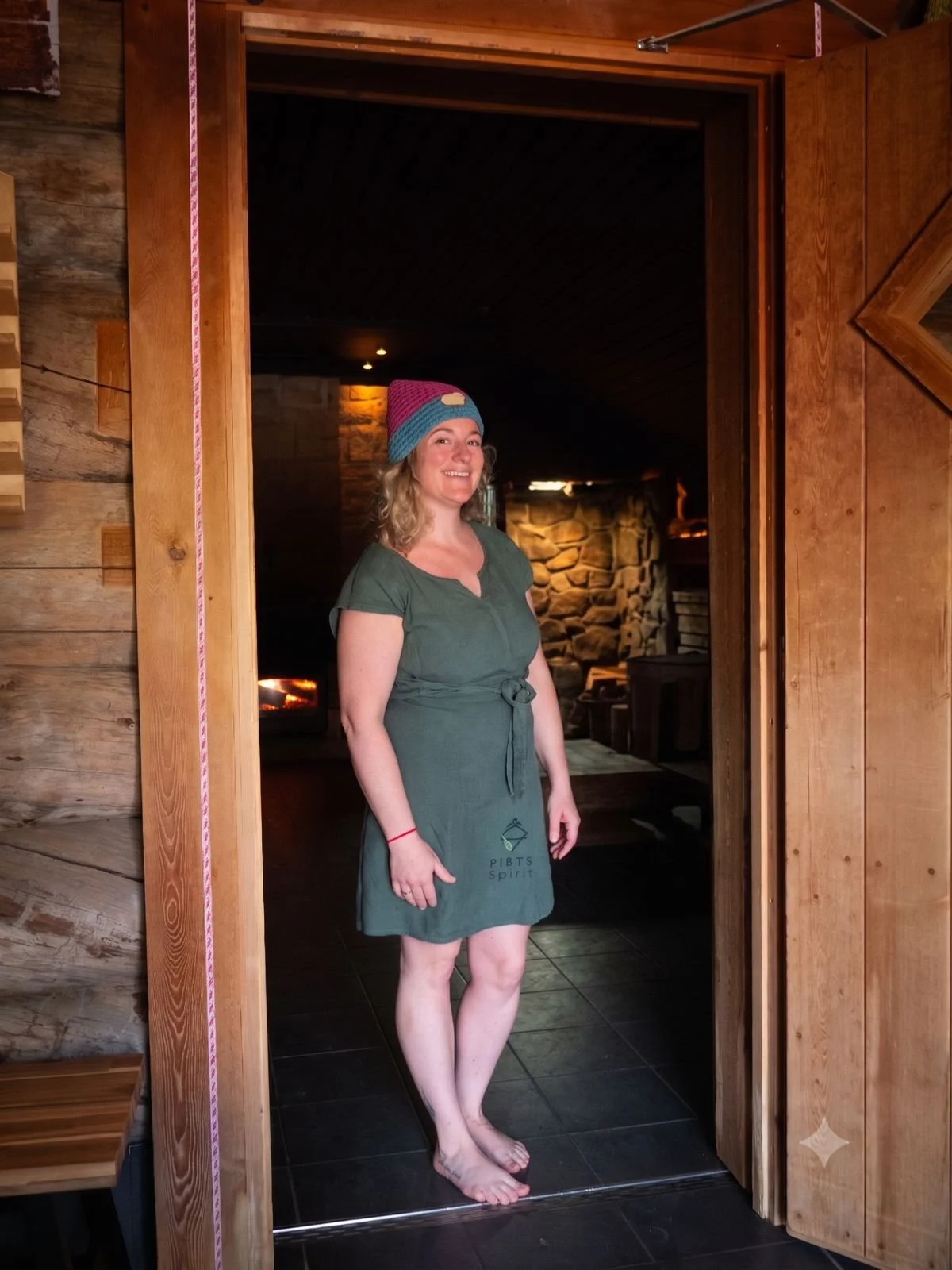 A woman standing in a doorway with wooden walls, smiling, wearing a green dress with a logo, and a colorful knit hat, inside a cozy, rustic room with stone fireplace.