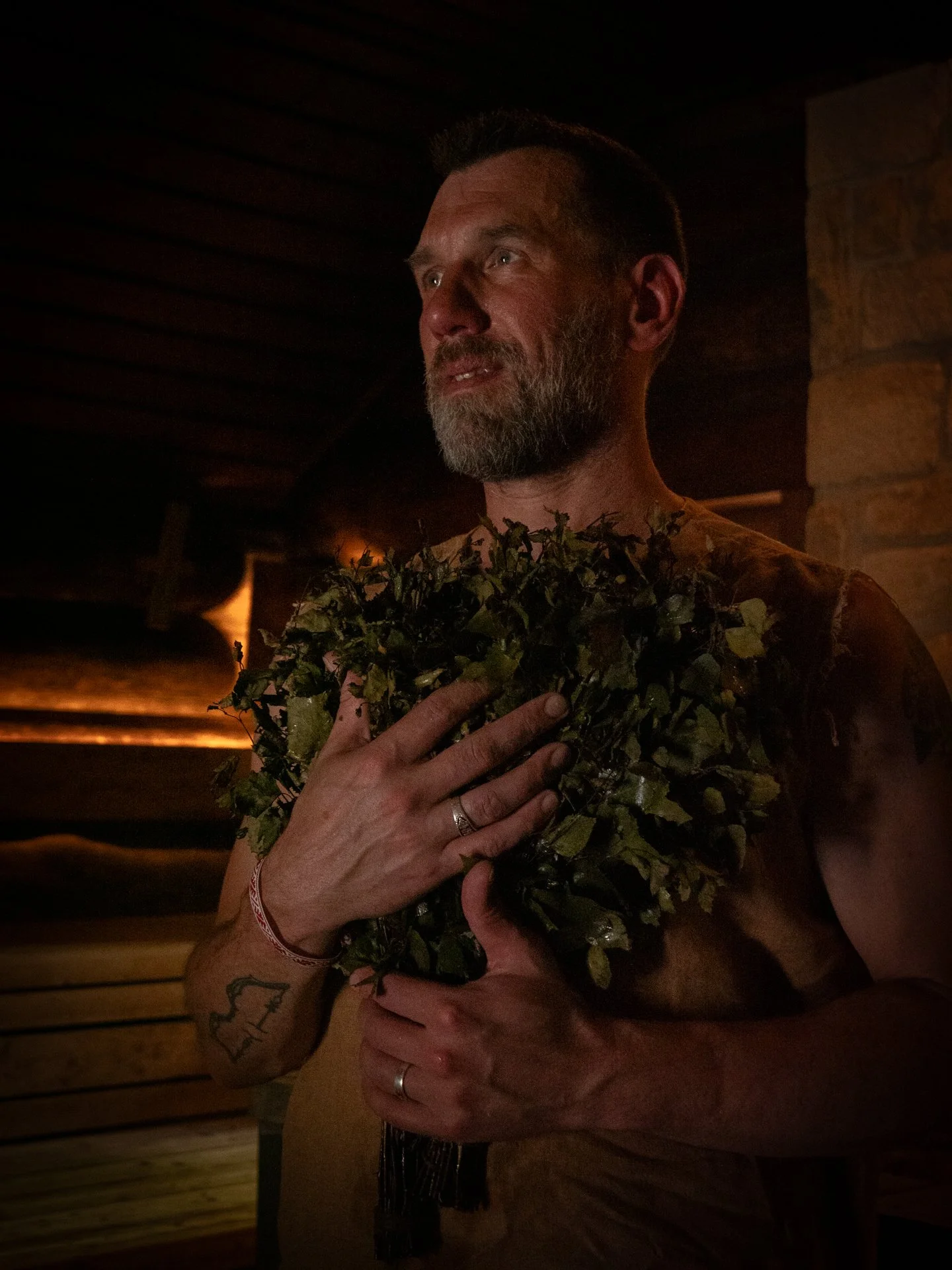 A bearded man with short hair holding a bunch of green leaves or herbs over his chest in a dimly lit room.