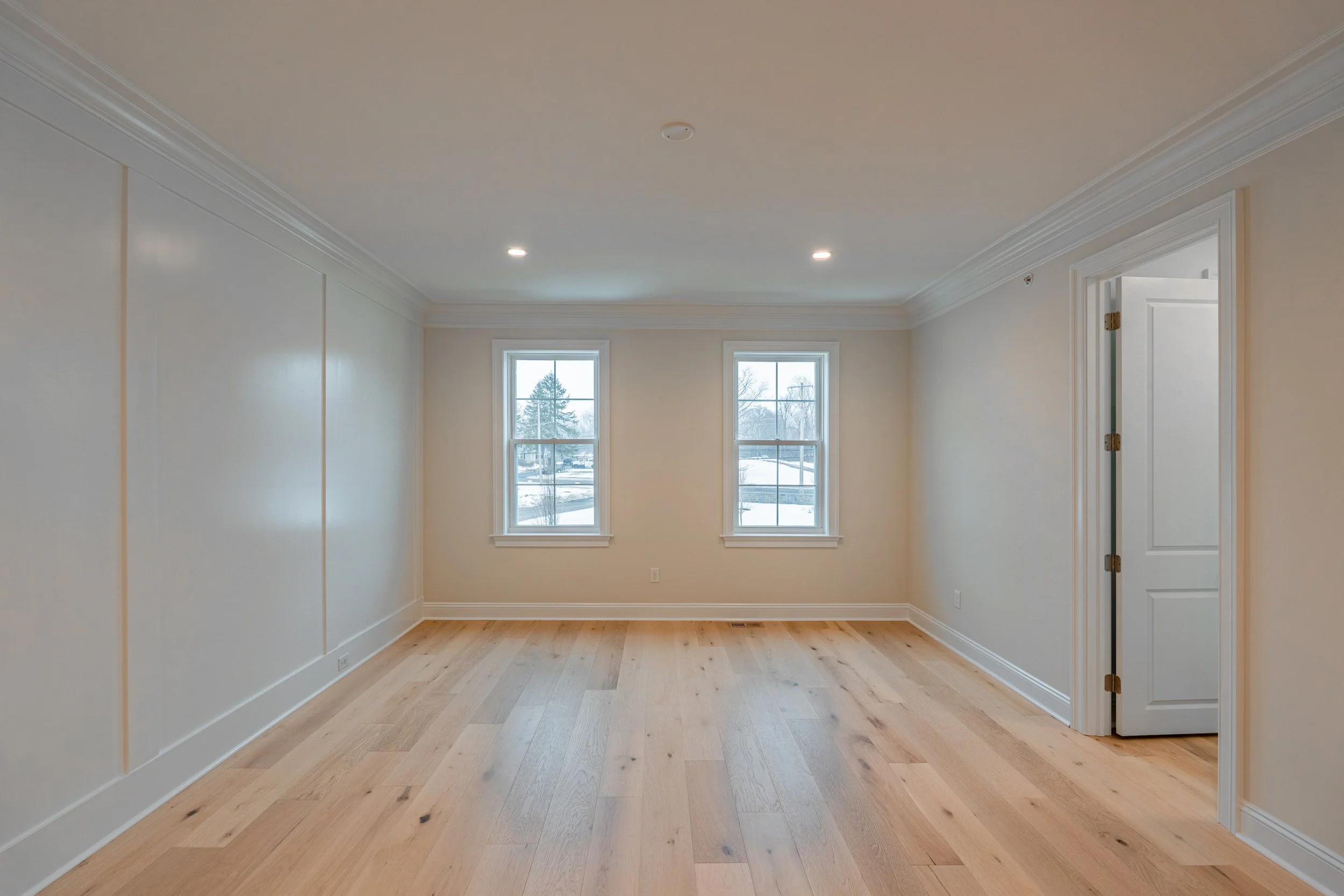 Empty room with light wood flooring, beige walls, crown molding, two windows, and an open door.