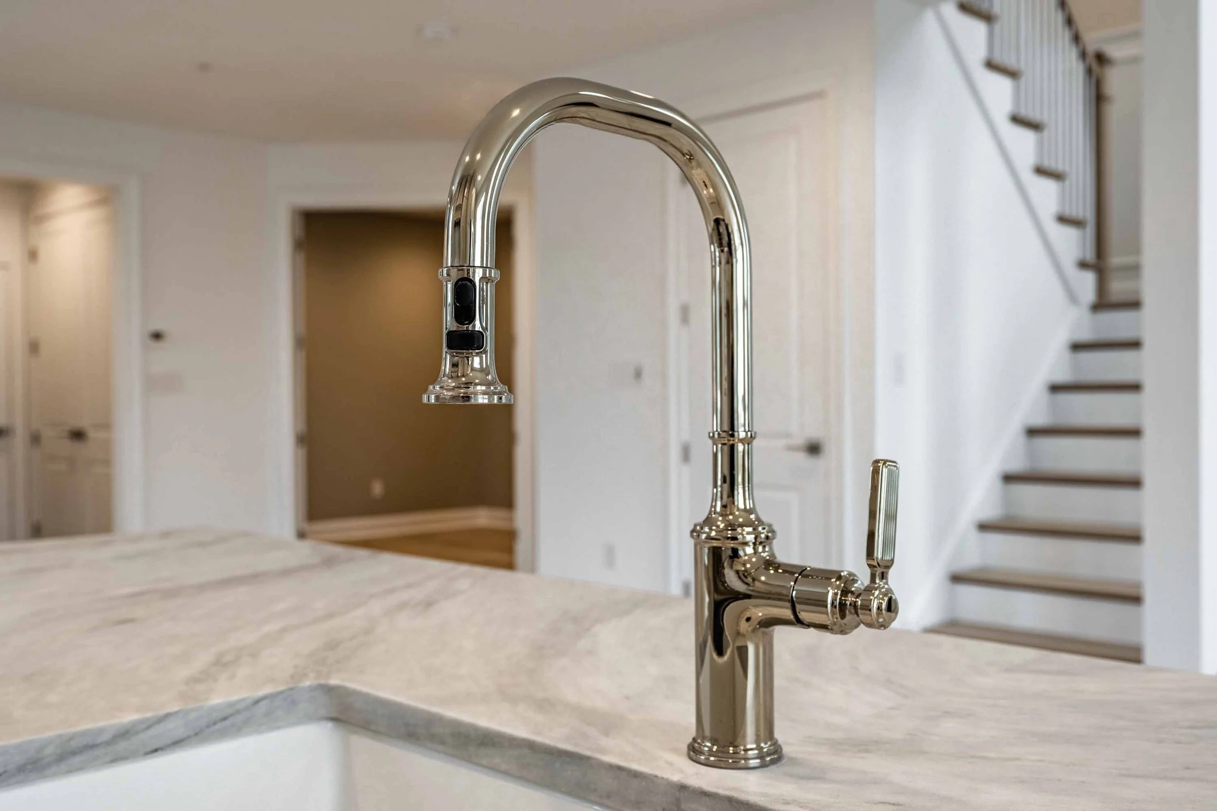 Close-up of a shiny, metallic kitchen faucet on a marble countertop in a modern kitchen with stairs in the background.