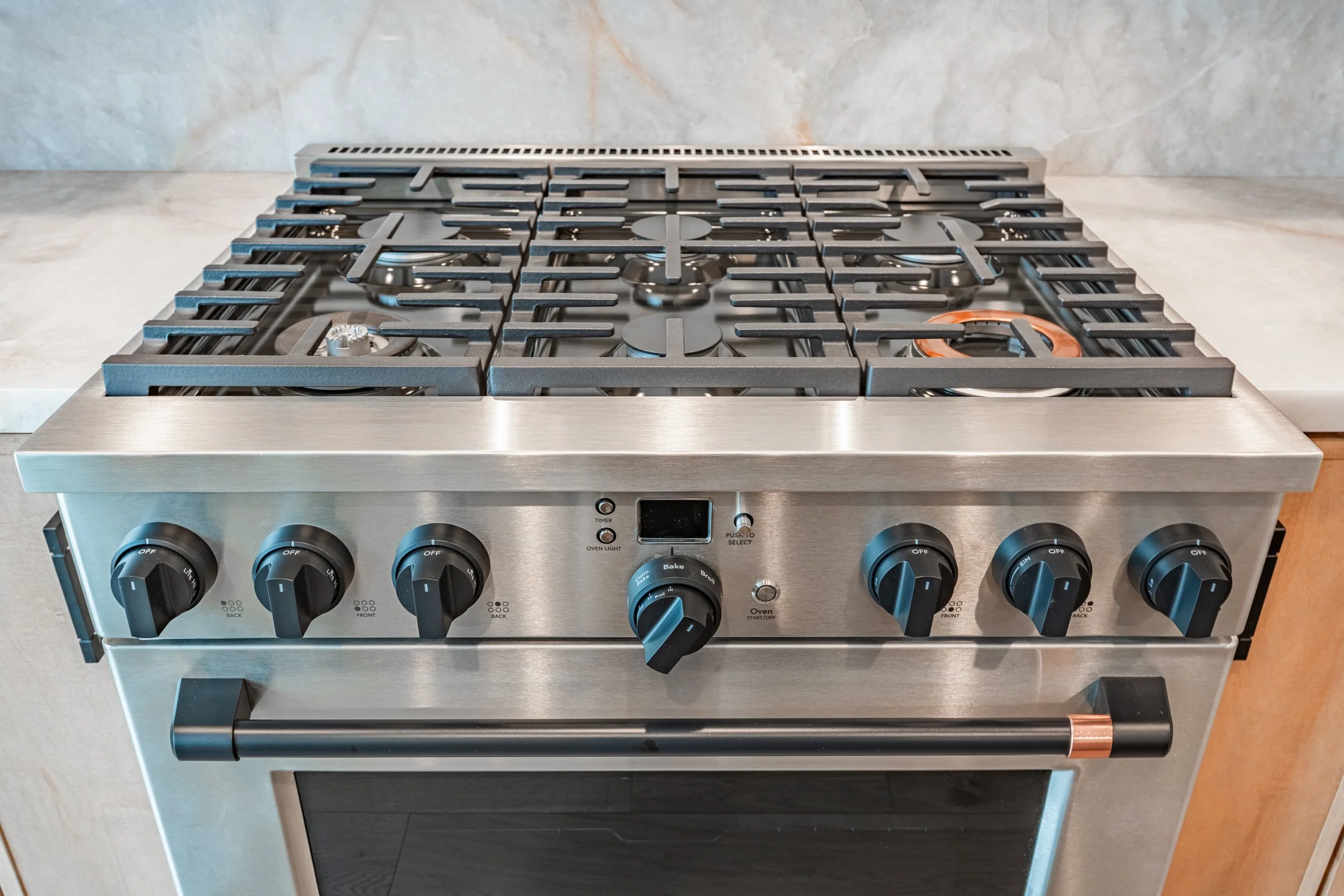 Stainless steel gas stove with black control knobs and grid burners, set on a kitchen counter with a marble backsplash.