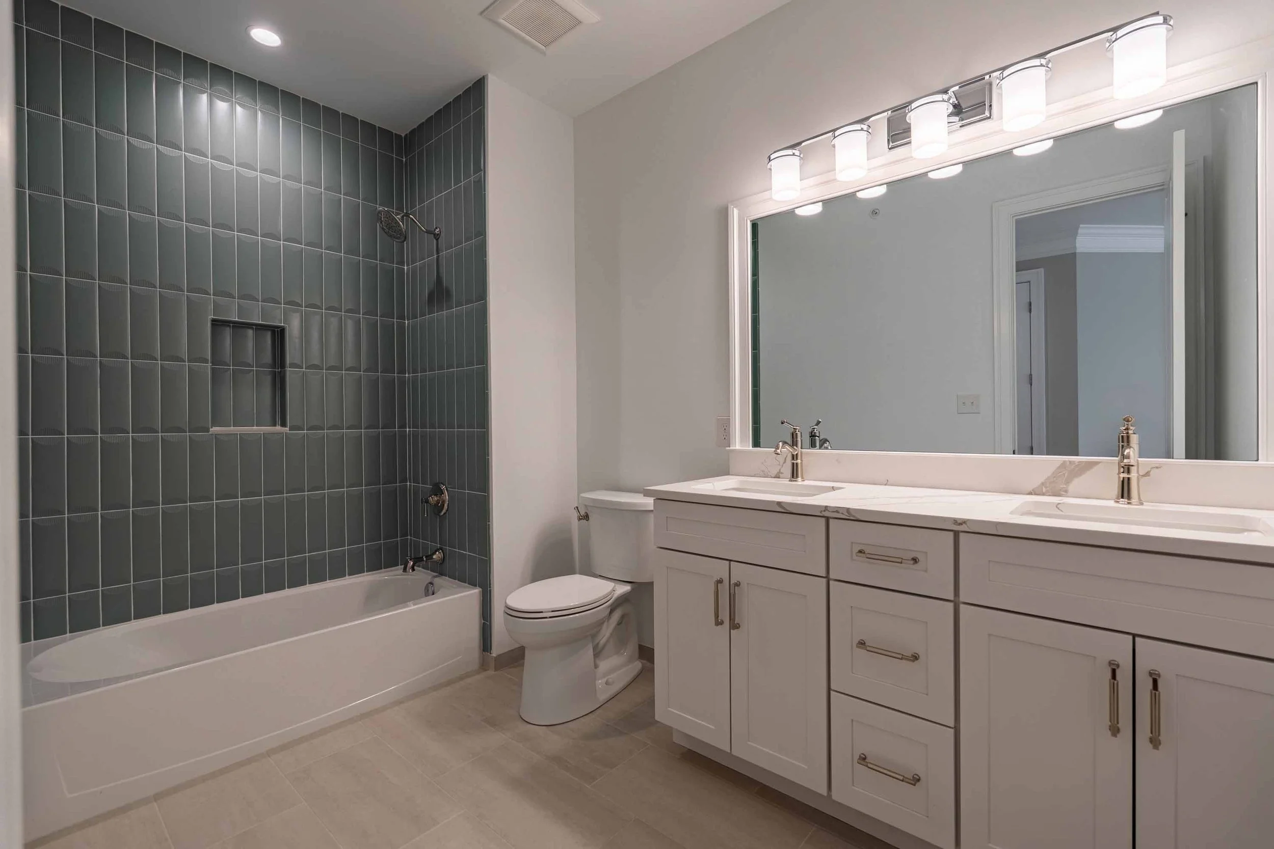 Bathroom with a bathtub and a shower area with dark gray tiles on the wall, a white vanity with drawers and gold hardware, a large mirror with a light fixture above, a toilet, and beige tile flooring.