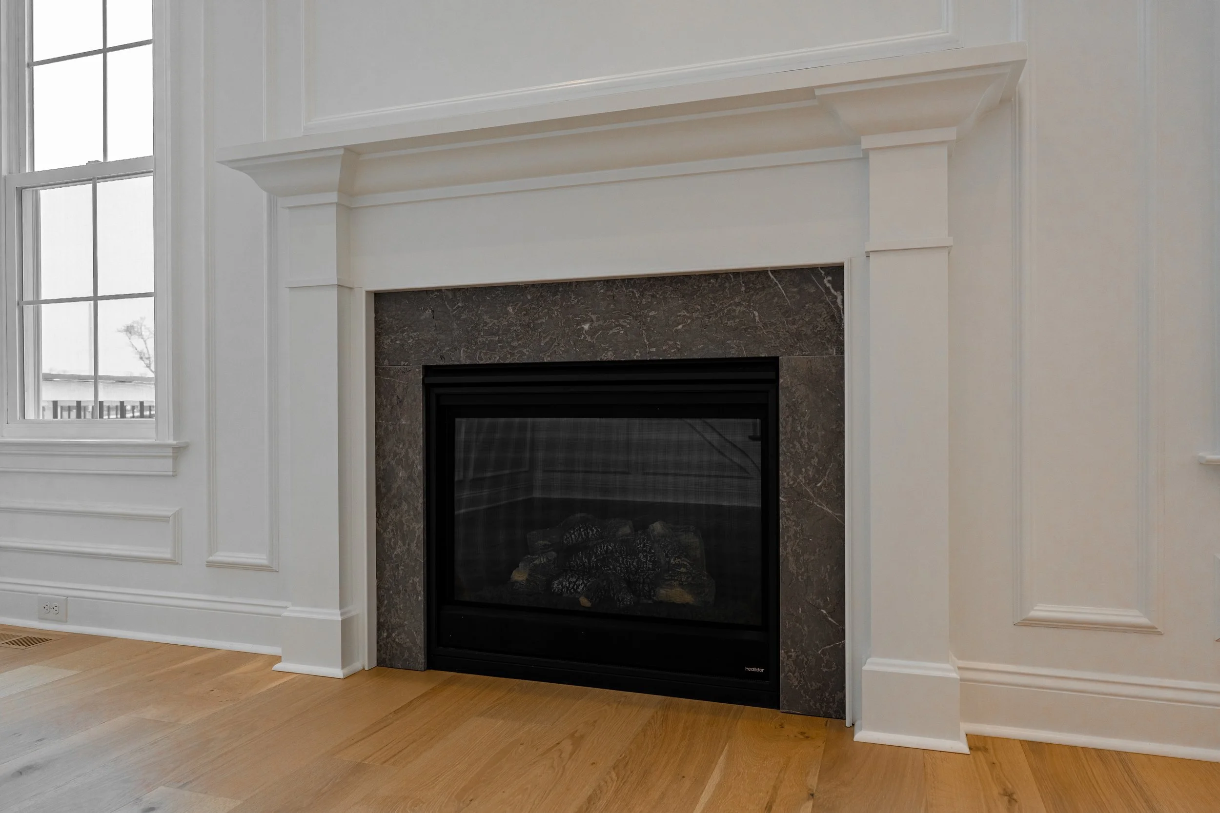 Interior view of a fireplace with a black frame and a marble surround in a white wall, next to a window with multiple panes, and hardwood flooring.