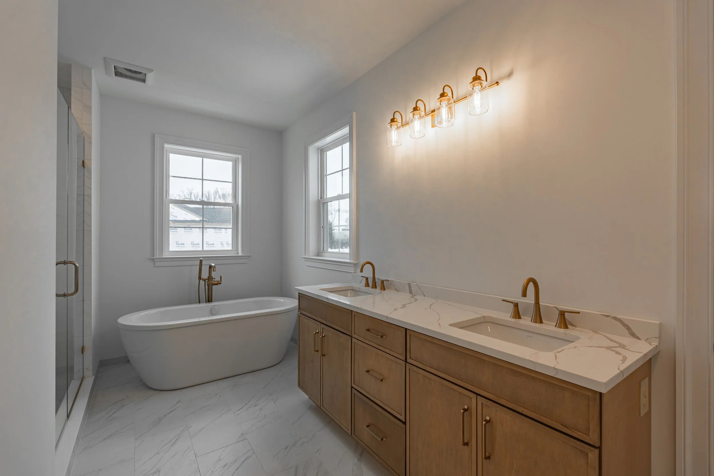 A bathroom with white walls and large windows, a freestanding bathtub, a wood vanity with a marble countertop, and a row of four vintage-style glass and brass light fixtures.