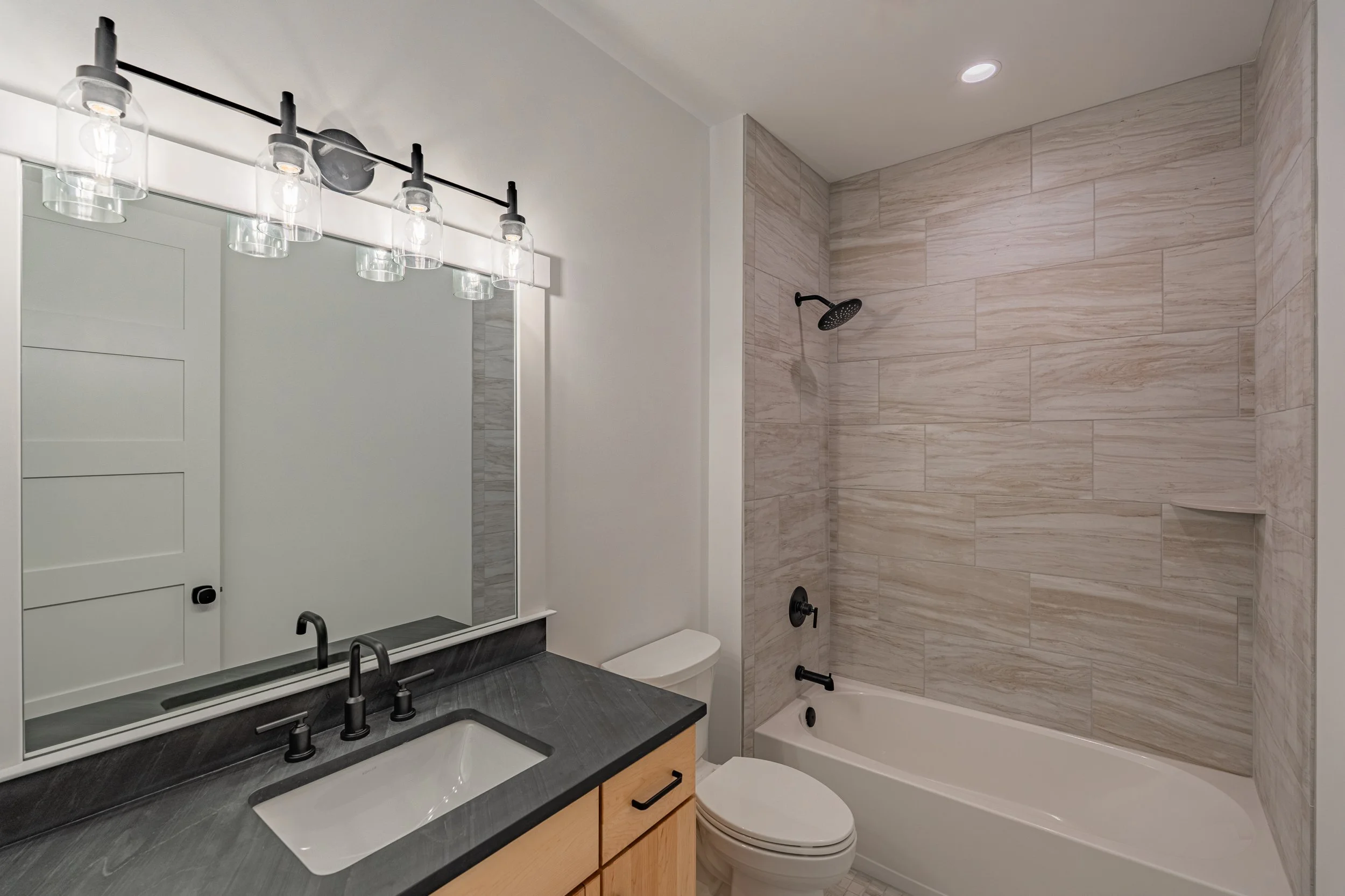 Modern bathroom with a black countertop sink, black faucets, a mirror with a white frame, a floating vanity, a toilet, and a bathtub with beige tiles on the wall.