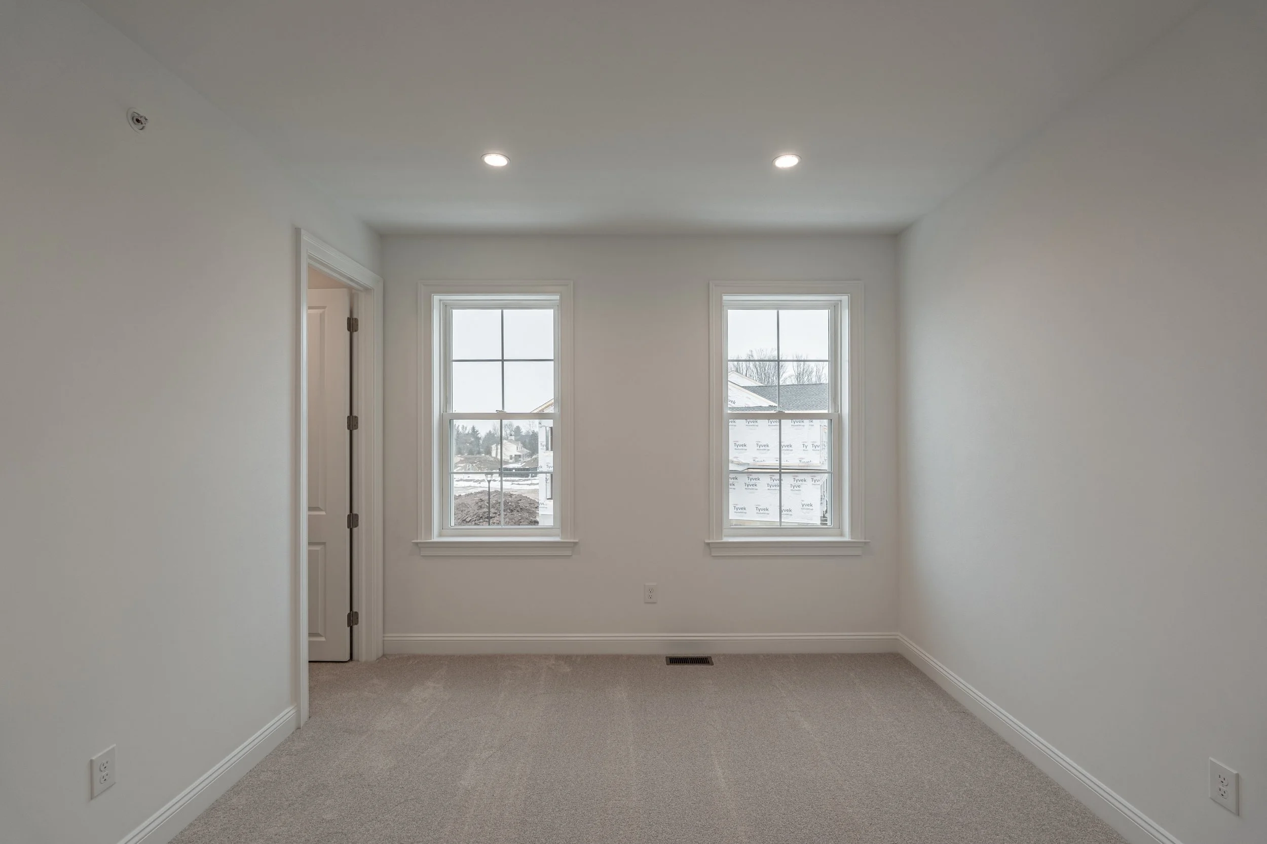 Empty room with two windows, white walls, beige carpet, and ceiling lights.