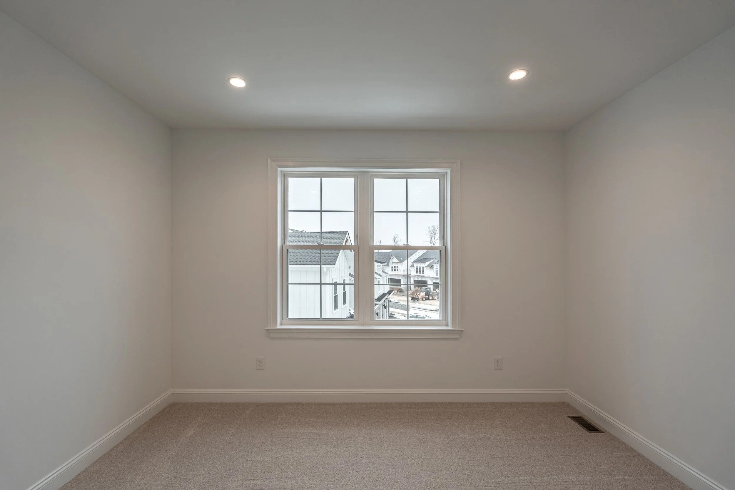 Empty room with beige carpet, white walls, and a window showing neighboring houses and a cloudy sky.