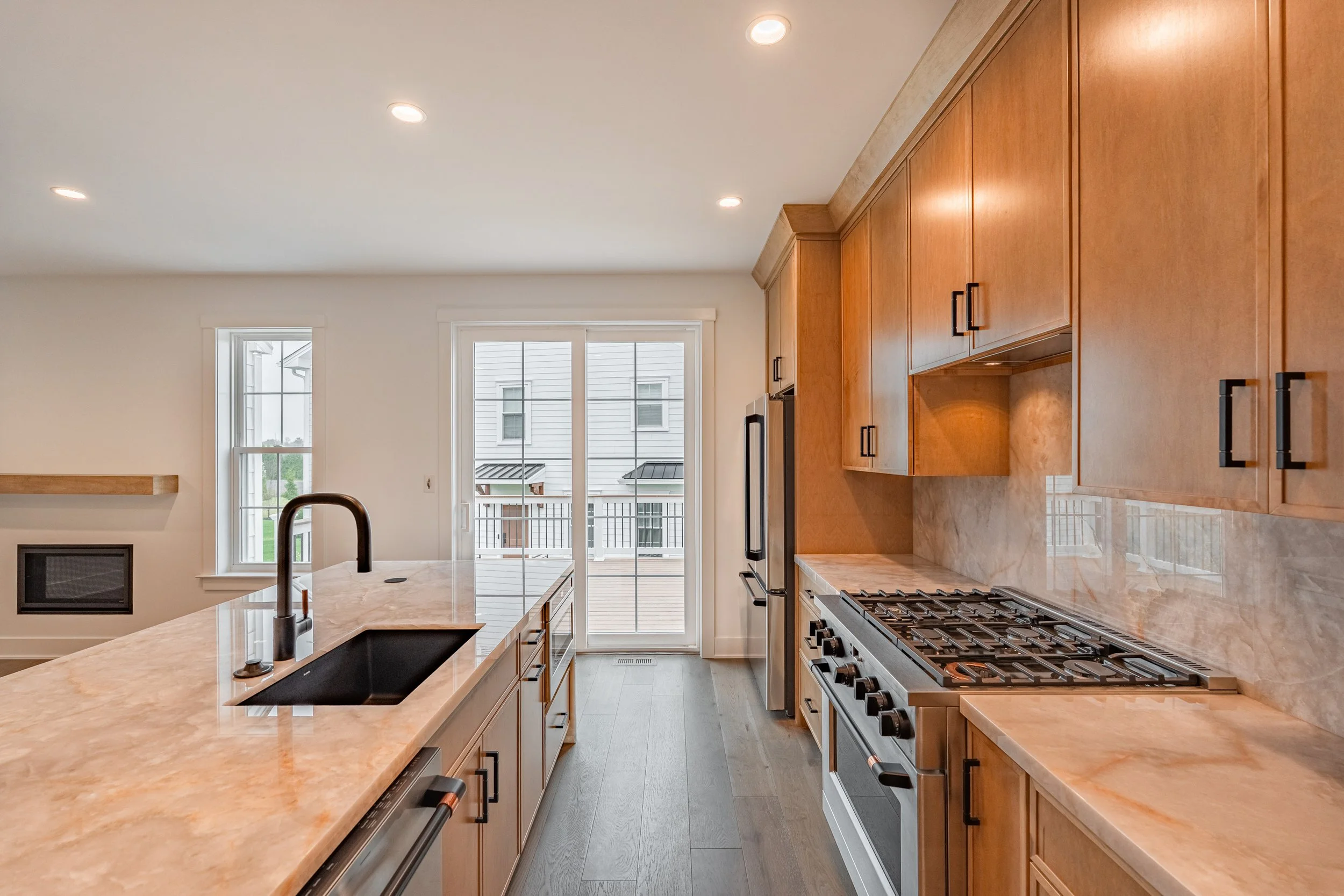 Modern kitchen with light wood cabinets, a marble countertop, black hardware, a black stove, a black sink with a high-arc faucet, and large windows with a sliding door leading to a balcony outside.
