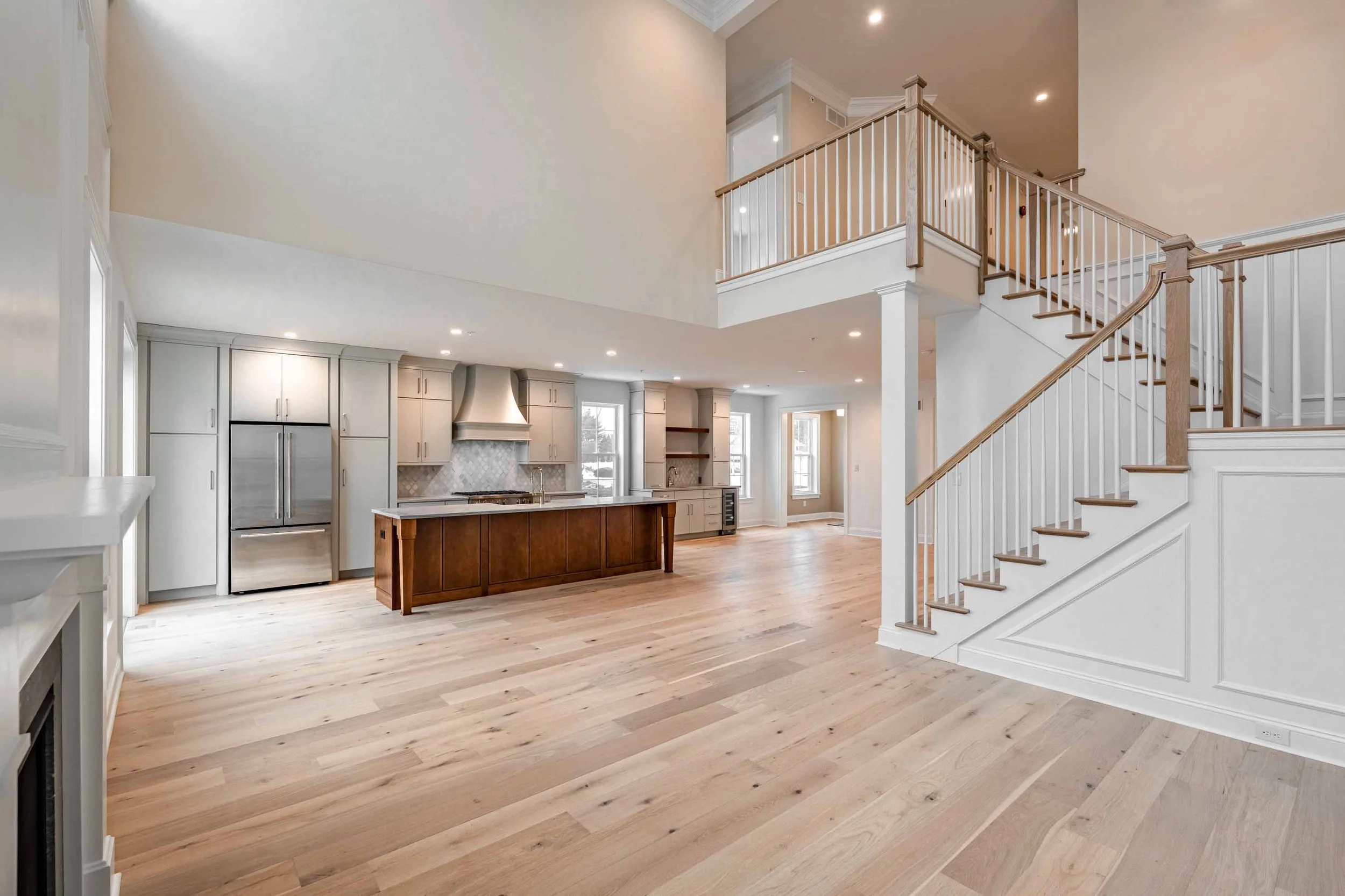 Open-concept kitchen and living space with light-colored hardwood floors, white walls, modern white cabinetry, granite countertops, stainless steel appliances, a staircase with wooden handrails, and high vaulted ceilings with recessed lighting.