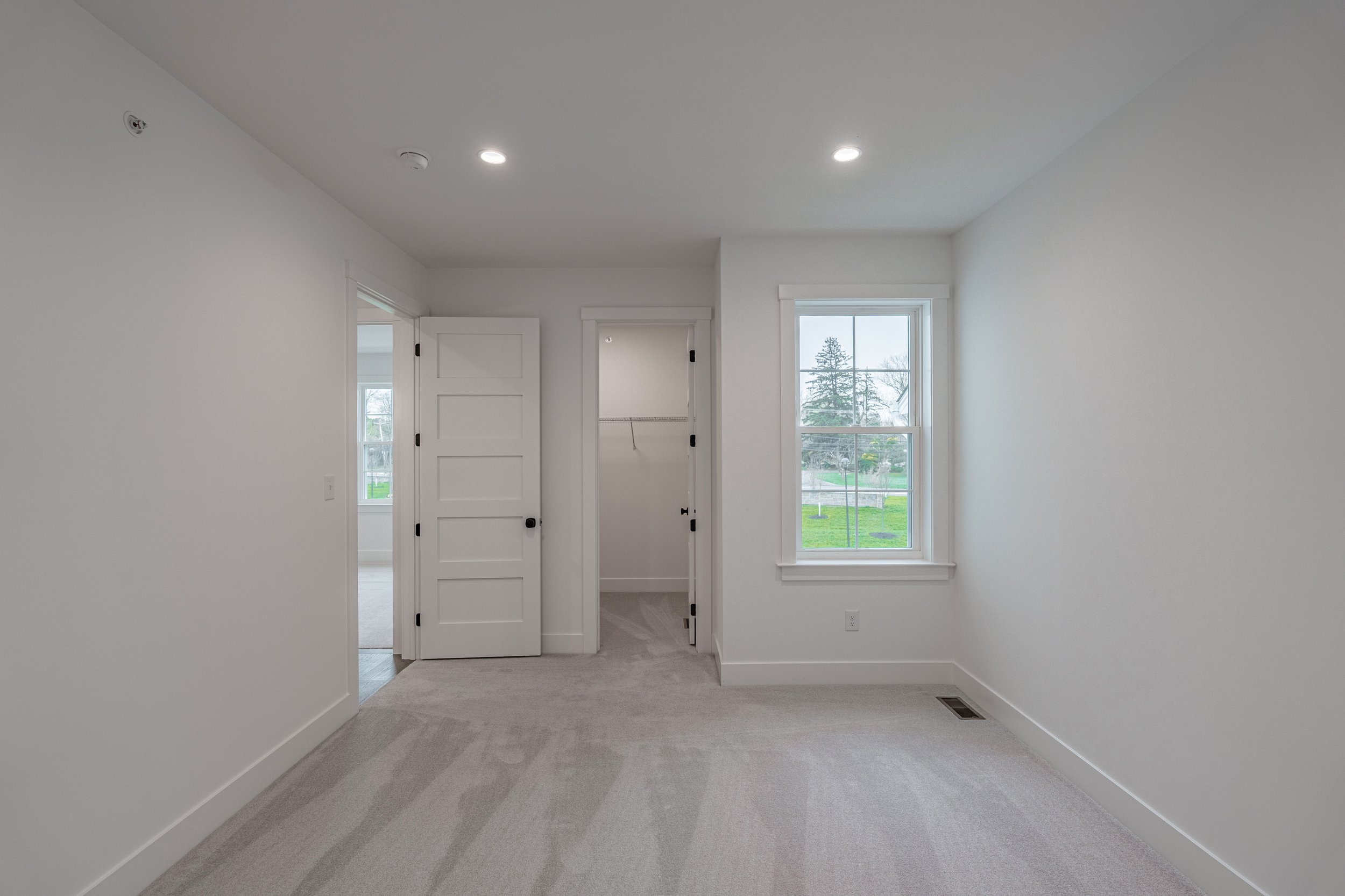 Empty room with white walls, two windows showing an outdoor view of trees and a lawn, and a small closet with double door. Carpeted floor, ceiling with recessed lights, and a floor vent.