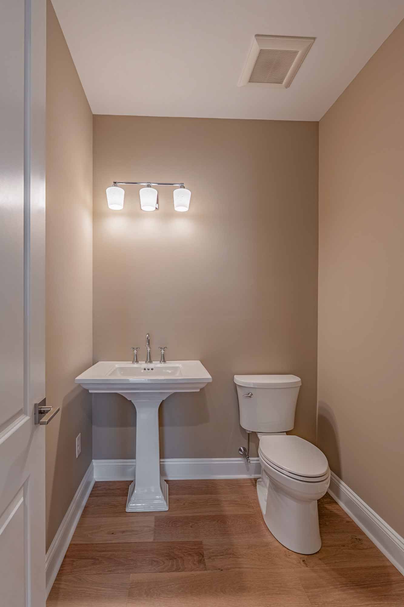 A small bathroom with beige walls, a white pedestal sink with a two-handle faucet, a toilet, a three-bulb wall sconce above the sink, a ceiling vent, and wood-style flooring.