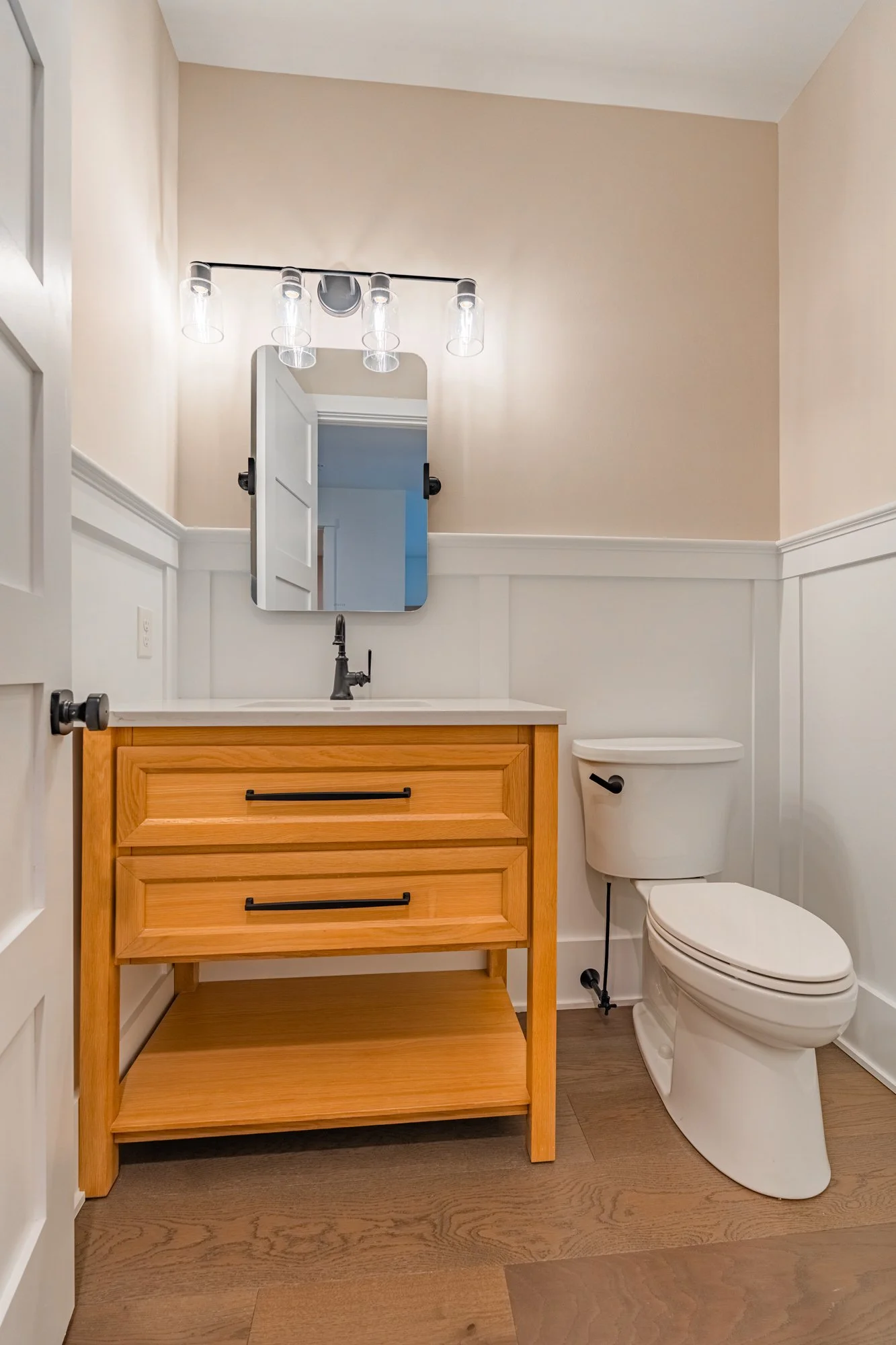 A modern bathroom with a white toilet, a wooden vanity with black handles, a small mirror, and a light fixture with four clear glass shades.