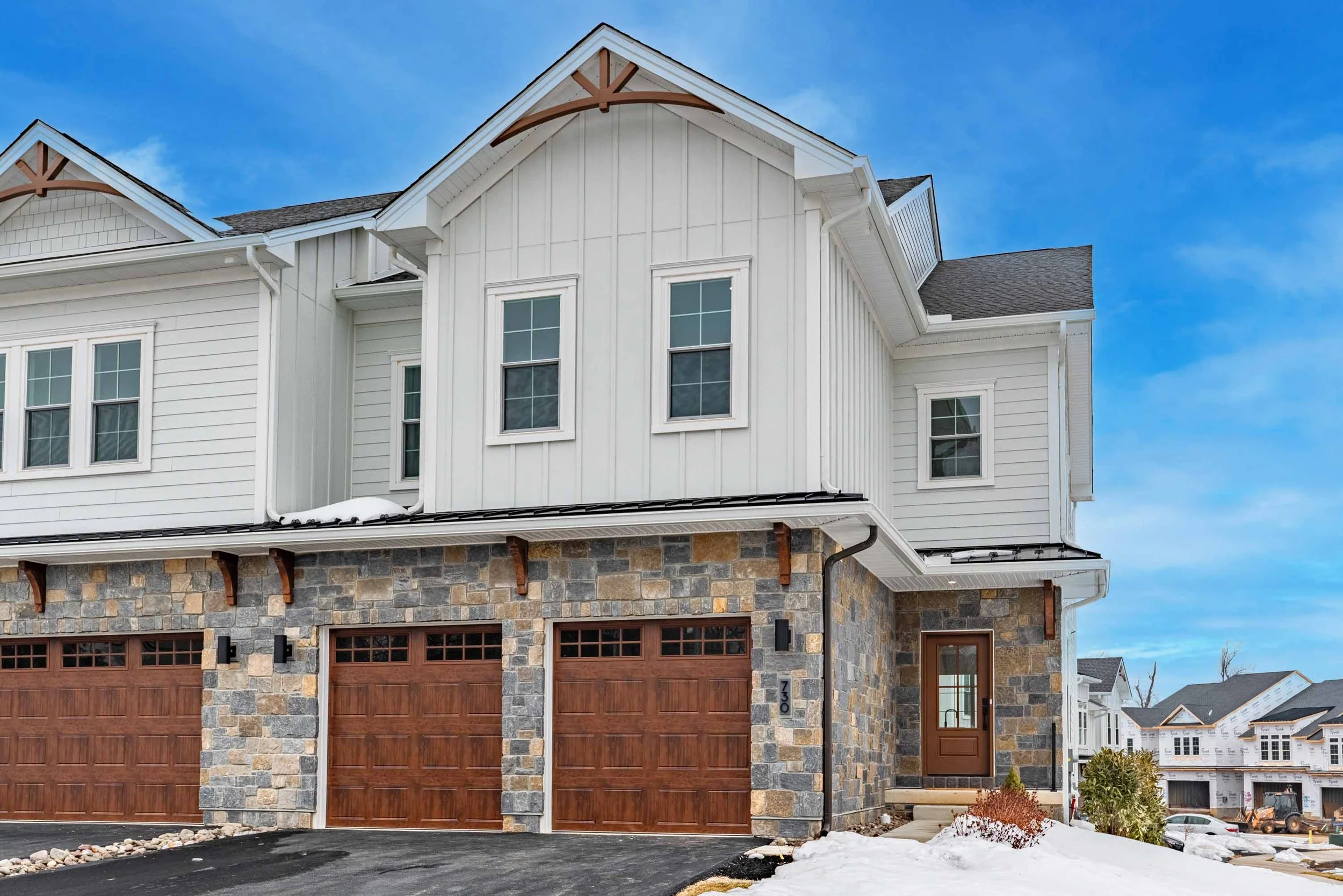 Front view of a newly constructed house with stone and wood garage doors, white siding, and multiple windows, under a blue sky with some snow on the ground.