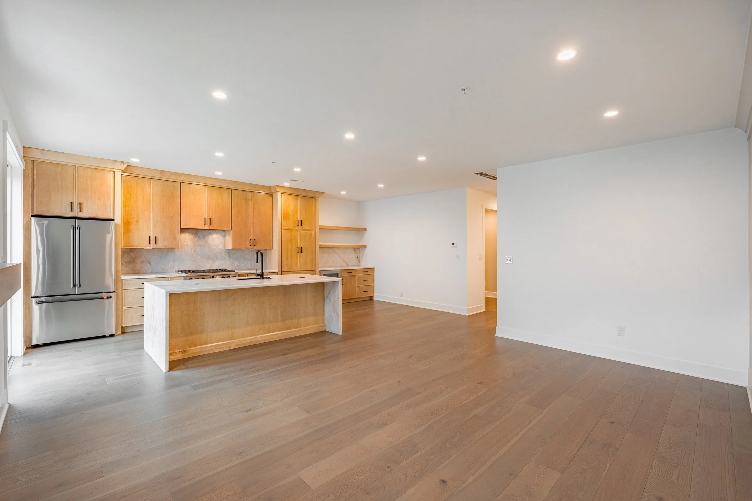 Modern kitchen with wooden cabinets, marble countertops, stainless steel appliances, and an open floor plan with wooden floors.