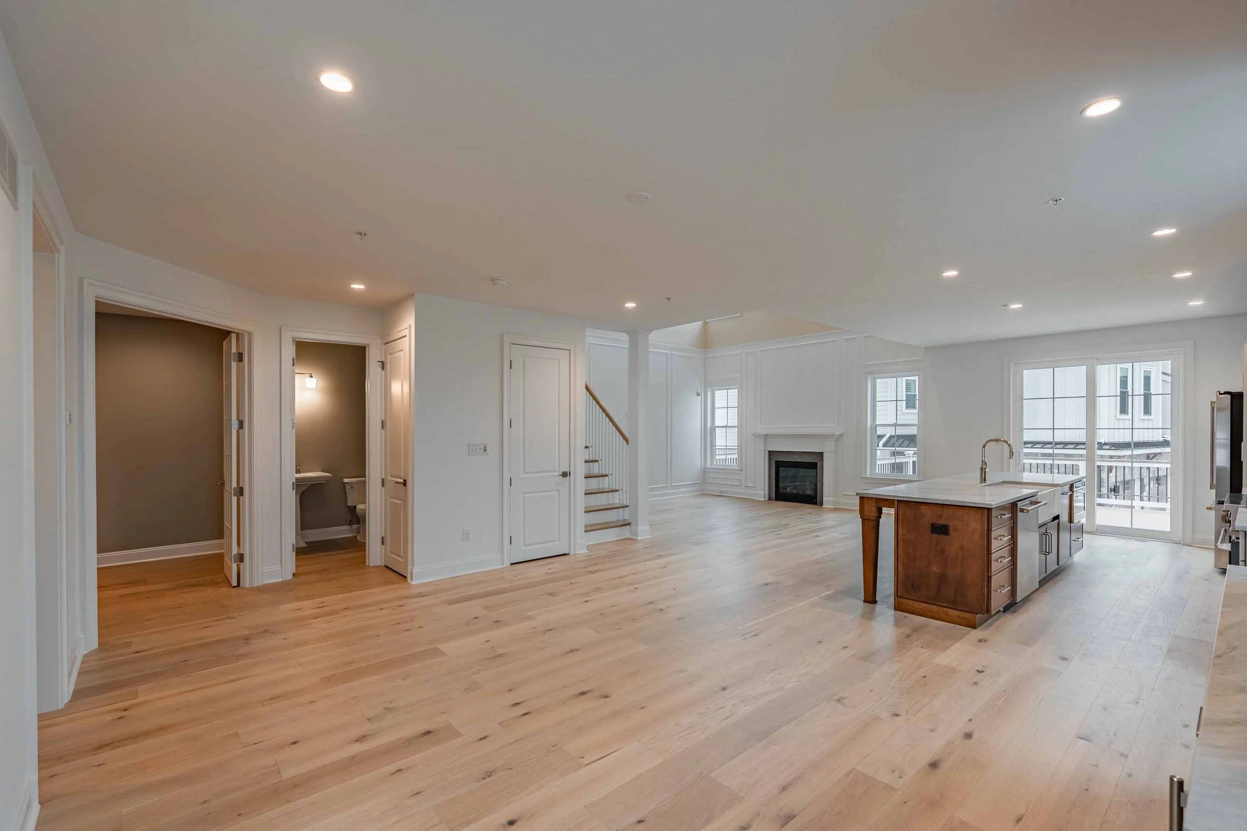 Spacious open-concept living room and kitchen area with hardwood floors, white walls, large windows, and a fireplace.