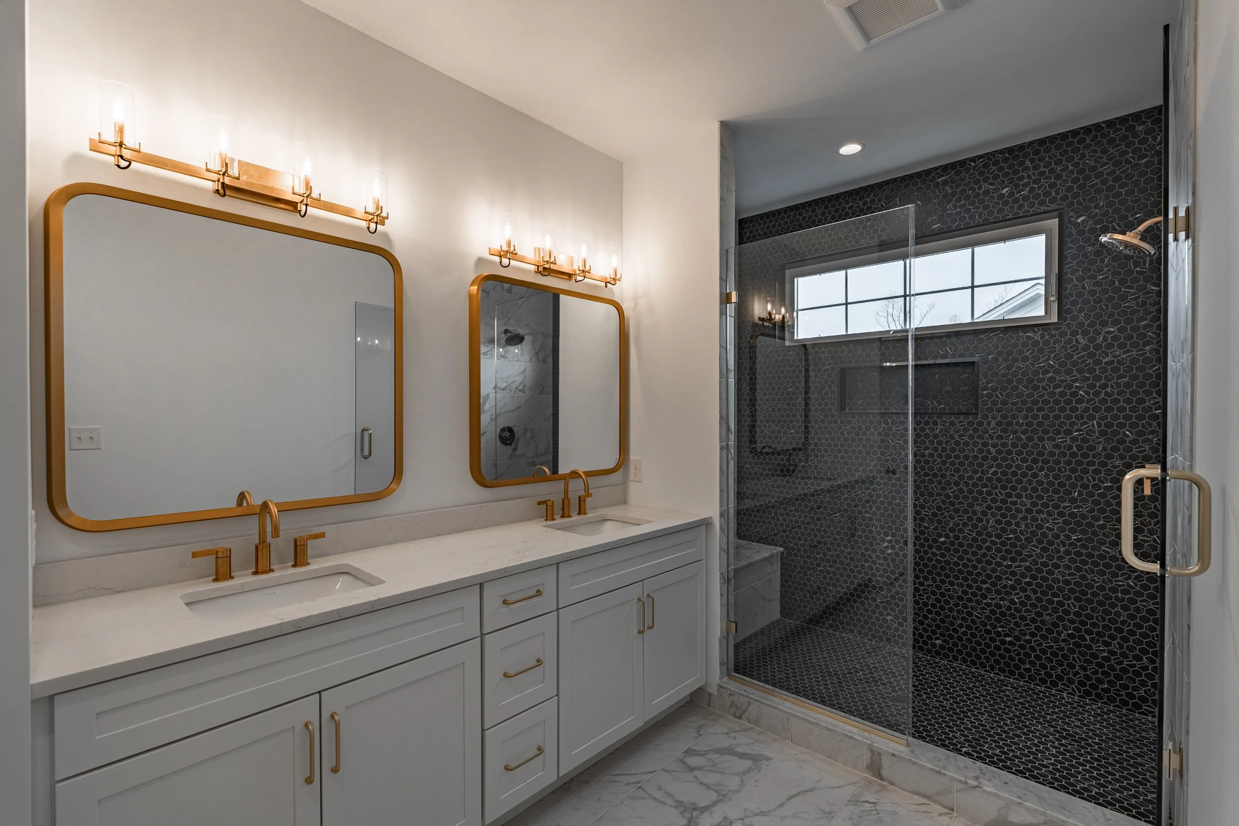 Modern bathroom with double vanity, gold fixtures, large mirrors, and a walk-in shower with black hexagonal tiles and a window.