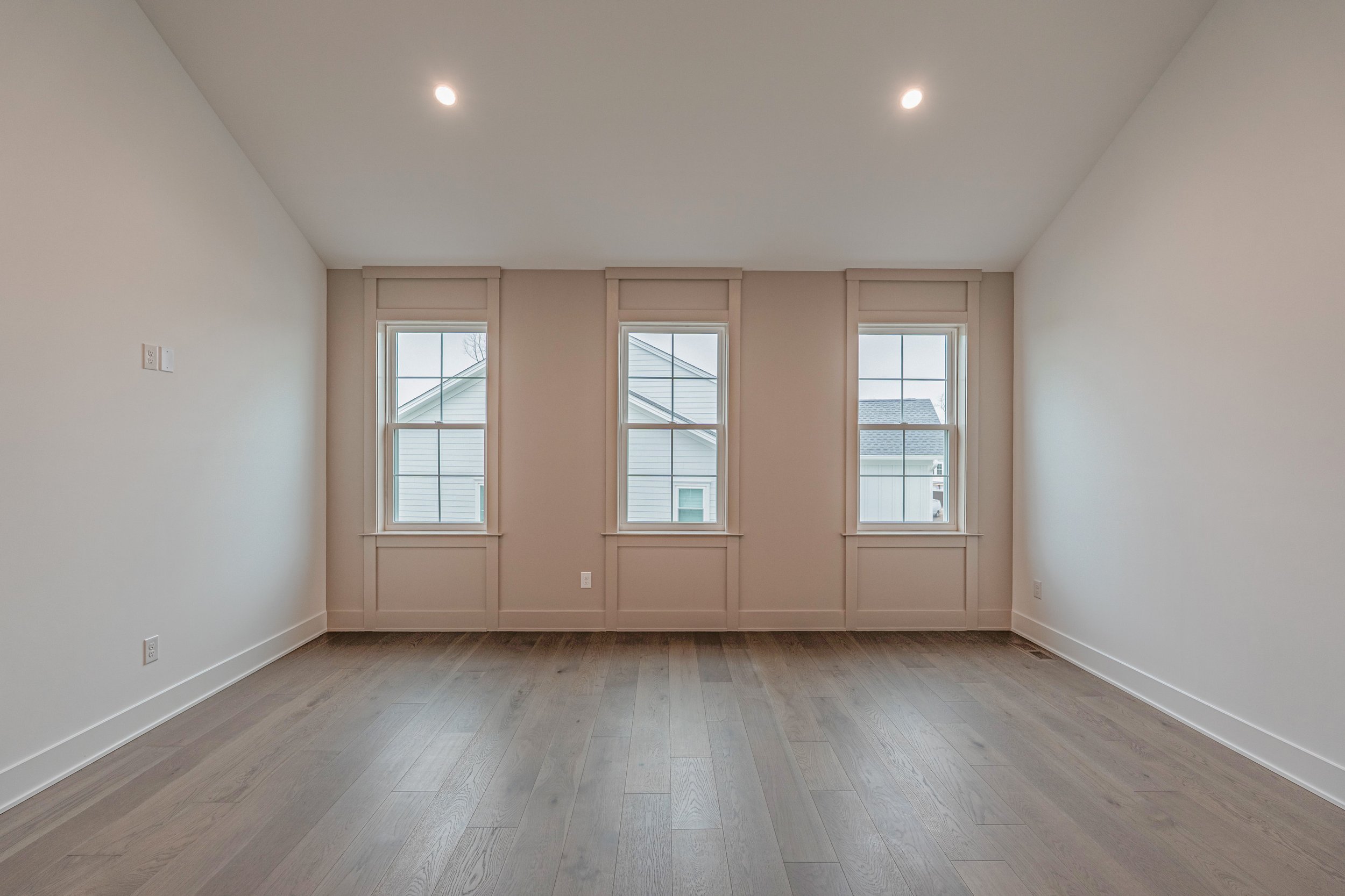 Empty room with three windows on the back wall, beige walls, white trim, white walls, and hardwood flooring.