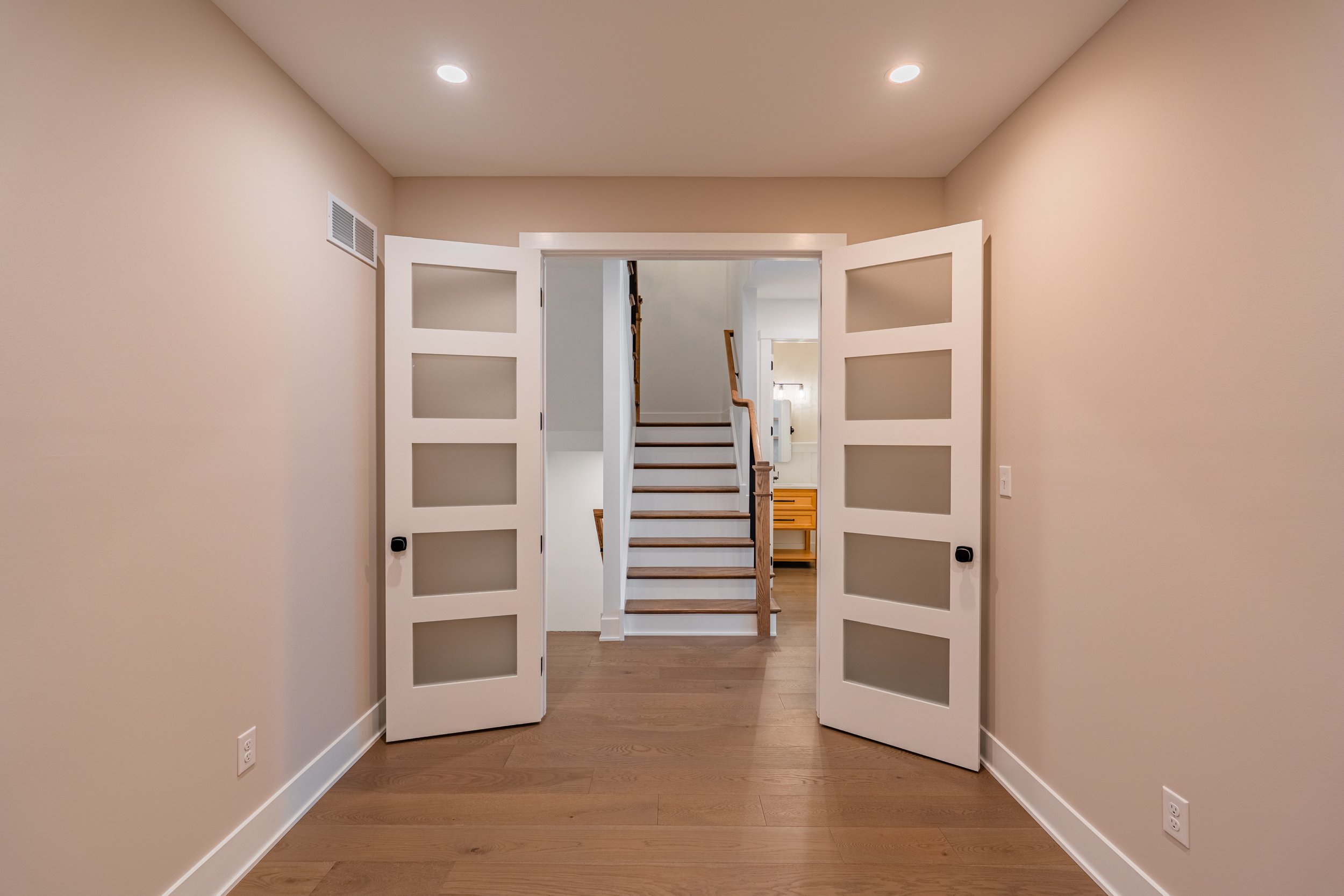 Interior view of a home doorway with separated white doors and a staircase visible through the opening, beige walls, wooden flooring, and ceiling lights.