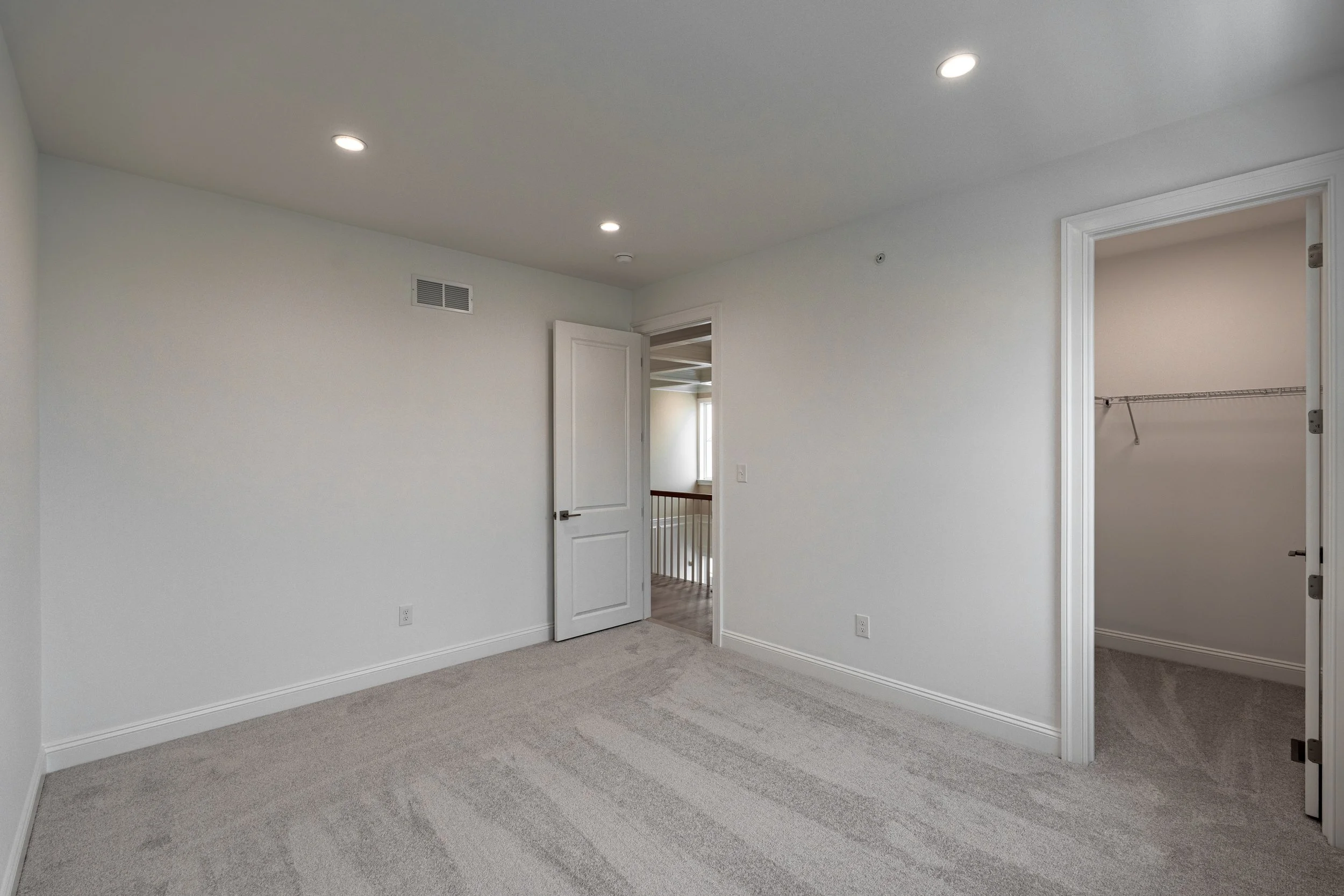 Empty bedroom with white walls, beige carpet, open closet, and a door leading to a hallway with a railing and window.
