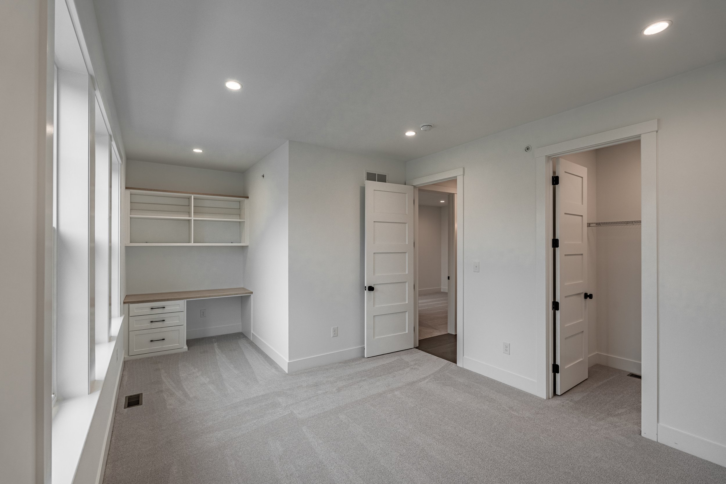 Empty room with white walls, ceiling with recessed lighting, and a beige carpet. There are built-in shelves and drawers on one wall, and open doors leading to other rooms or closets.