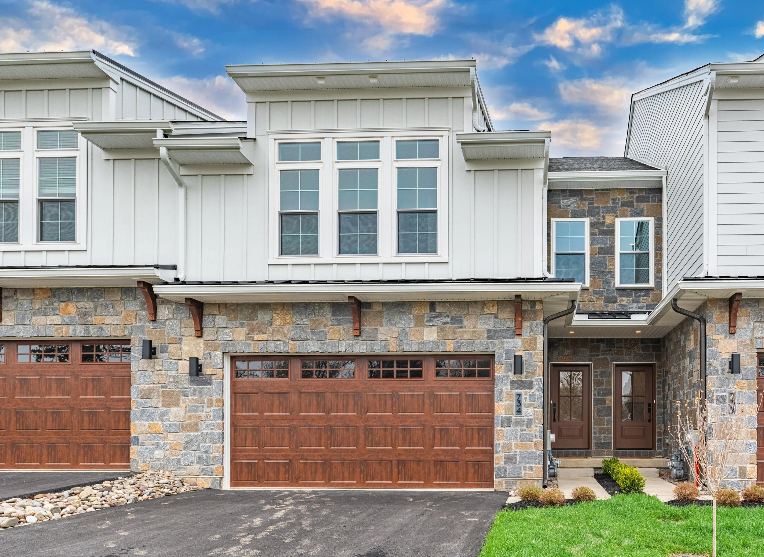 Modern house with stone and white siding exterior, four-car garage with wooden doors, and front yard with green grass and small bushes.