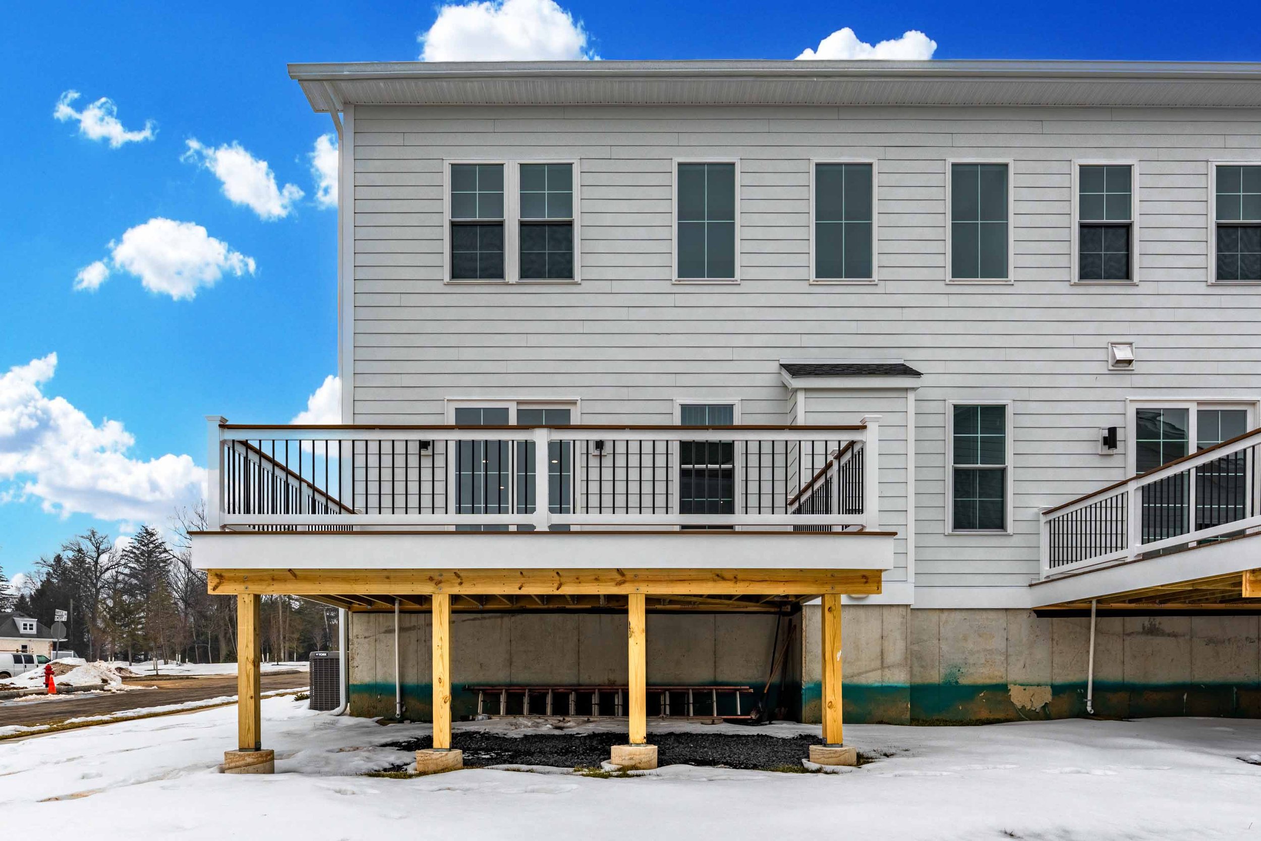 A house under construction with a white exterior, multiple windows, and a deck area with railings, set against a blue sky with clouds. The ground is covered with snow, and an unfinished porch with wooden support beams is visible at the bottom.