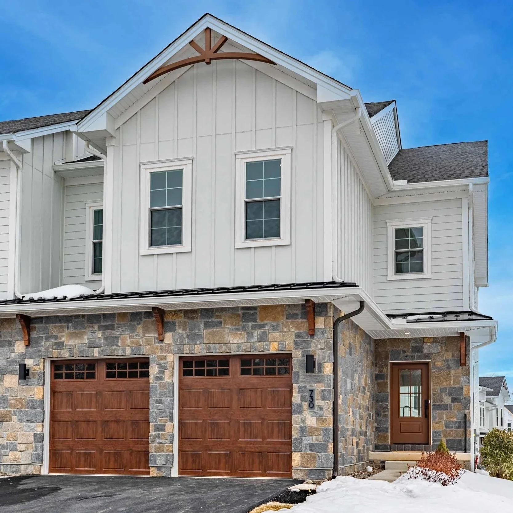 A modern two-story house with a stone facade, wooden garage doors, and white siding above. The house has three small windows on the second floor, a front door with a small window, and a garage with two doors. Snow is on the ground and the sky is clear and blue.
