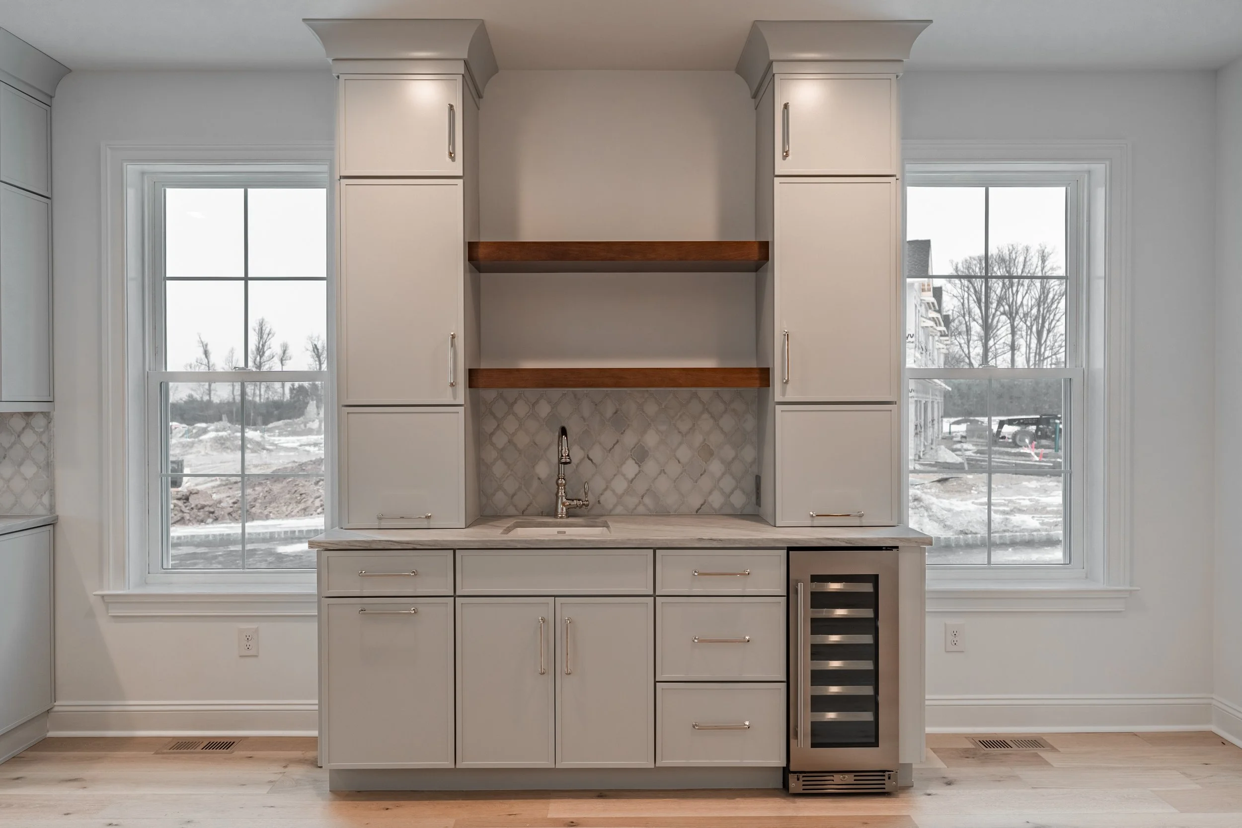 Kitchen with white cabinets, two large windows, a marble countertop, and built-in appliances, with a view of a snowy outdoor scene.