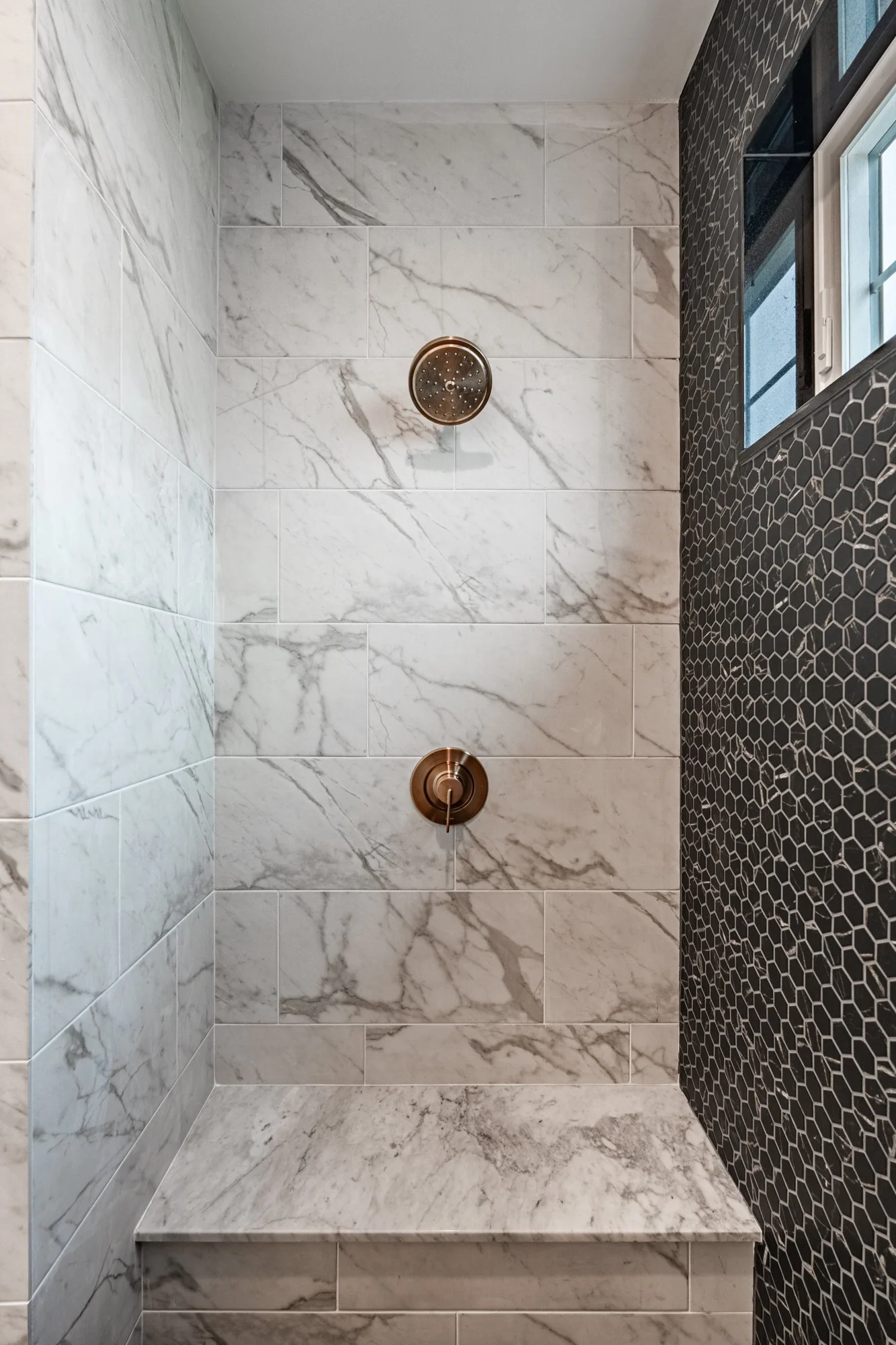 View of a modern shower with white marble tile walls, a built-in marble bench, a bronze showerhead, and a black honeycomb tile accent wall next to a window.