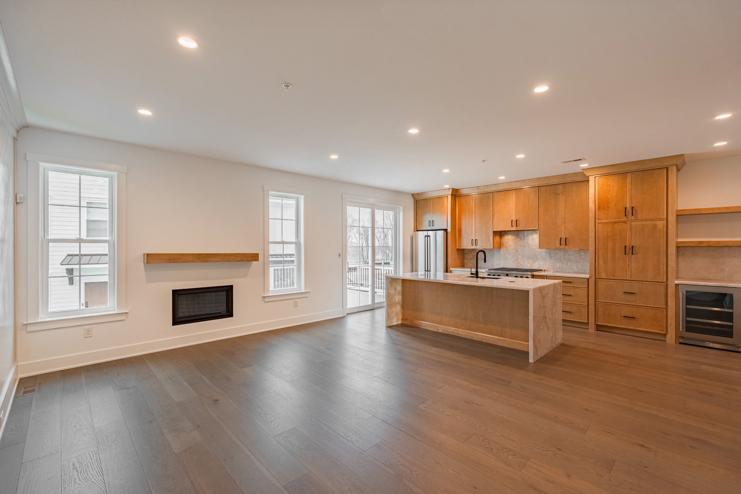 Interior view of a modern kitchen and living area with hardwood floors, white walls, multiple windows, a fireplace, wooden cabinets, and a kitchen island with a sink.