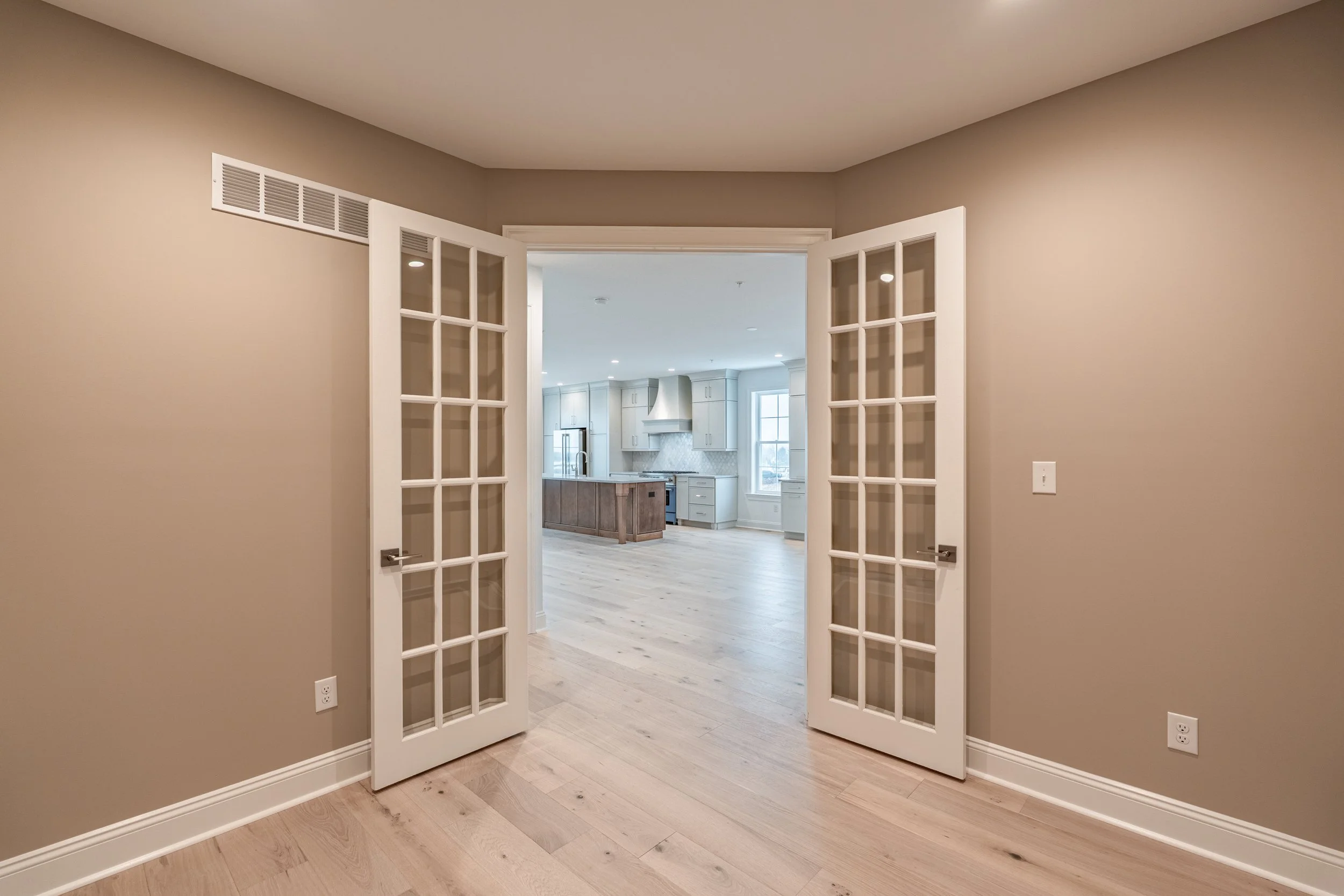Interior view of a house with French doors leading to a kitchen with white cabinets, a center island, and a window, with light hardwood floors and neutral walls.