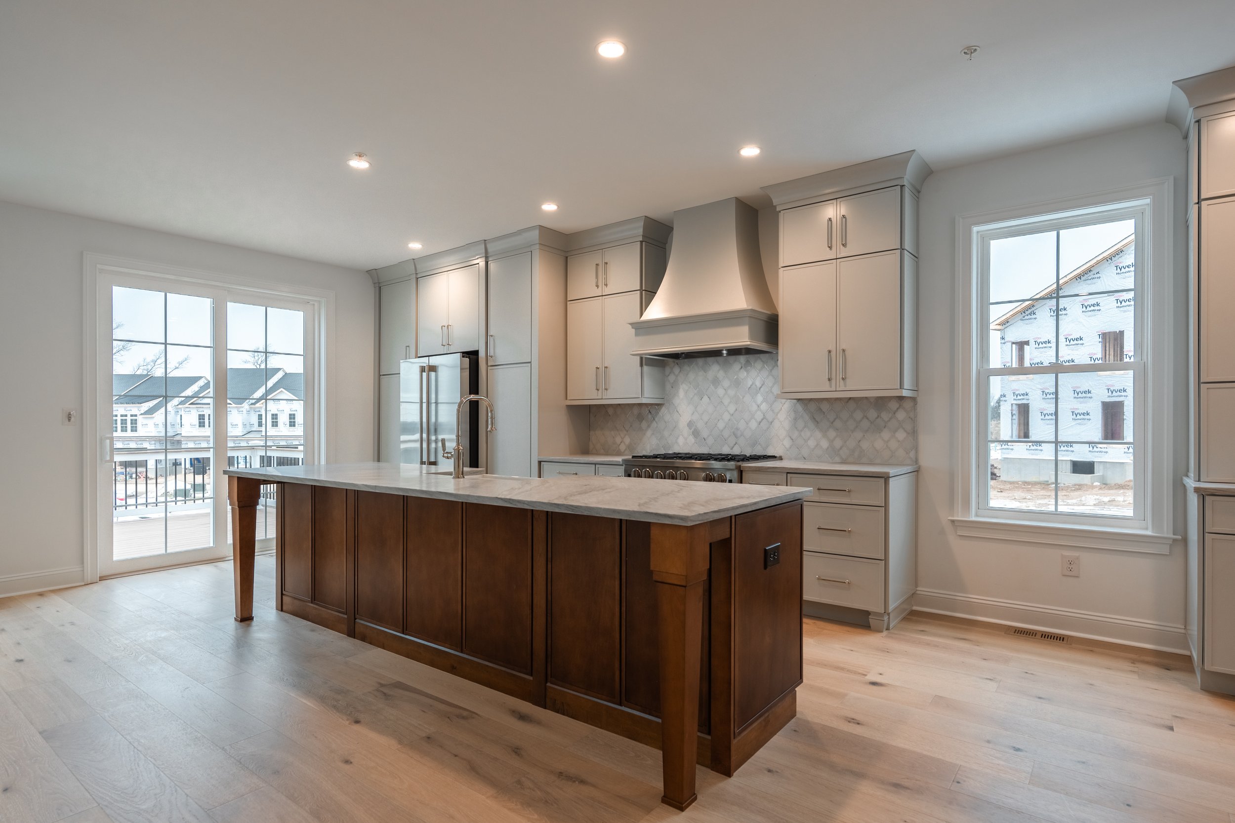 Modern kitchen with white cabinets, stainless steel appliances, a marble-topped island, and large windows showing neighboring houses under construction.