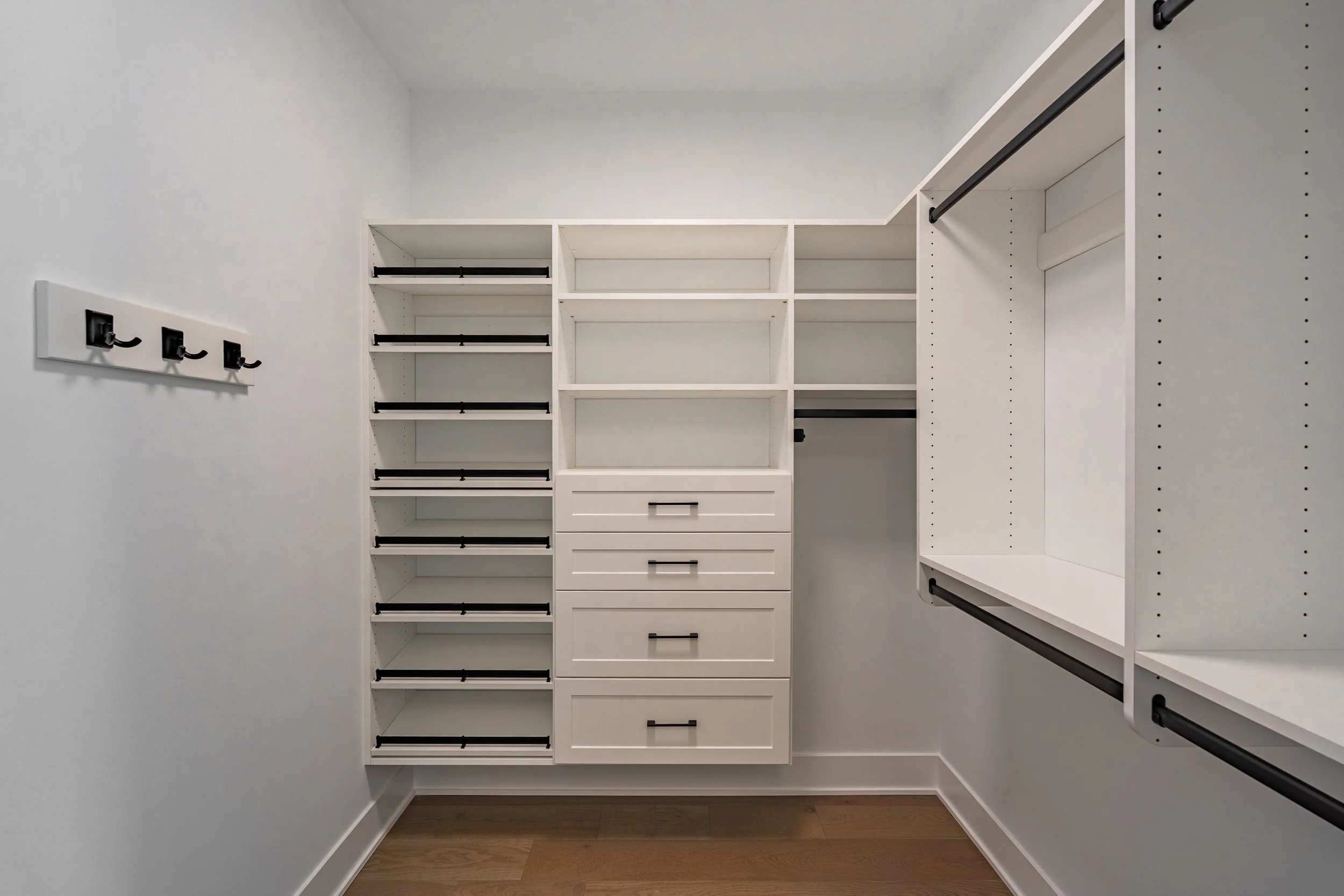 Empty walk-in closet with white shelving units, drawers, and black hooks, featuring a rod for hanging clothes on the right side.