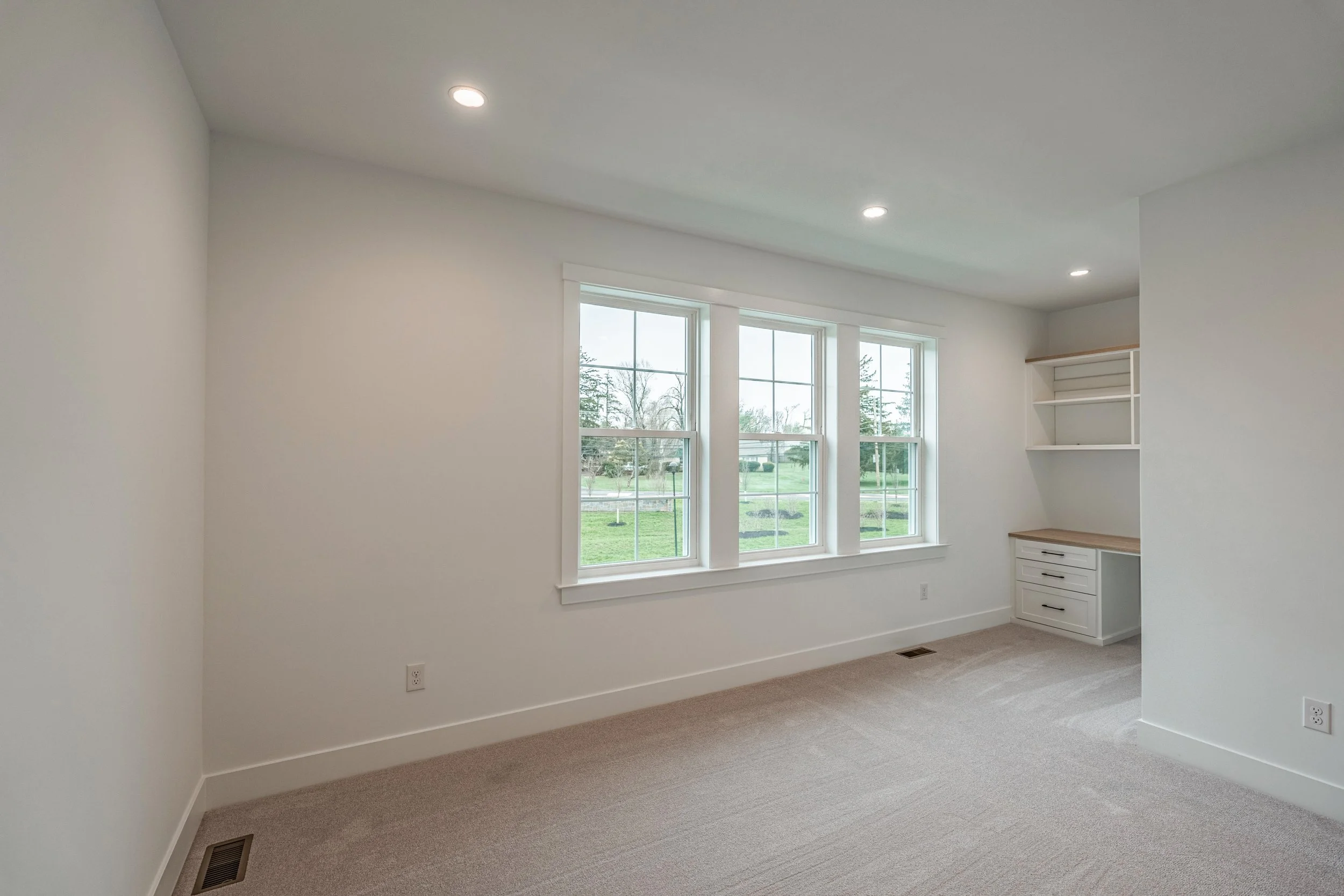 The image shows an empty room with white walls, a beige carpet, three large windows allowing natural light, and built-in white shelves on the right wall. There are ceiling lights and an electrical outlet near the floor.