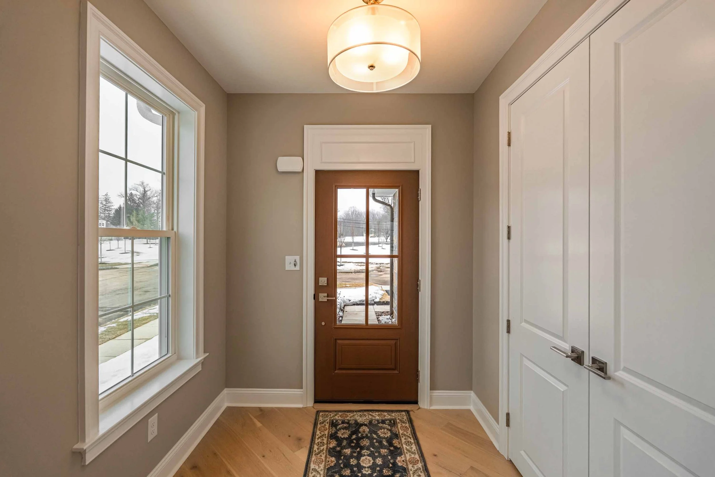 A hallway in a house featuring a wooden front door with glass panels, a white window on the left, a white storage cabinet on the right, beige walls, hardwood flooring, and a patterned area rug in front of the door. Exterior view shows snow and leafle