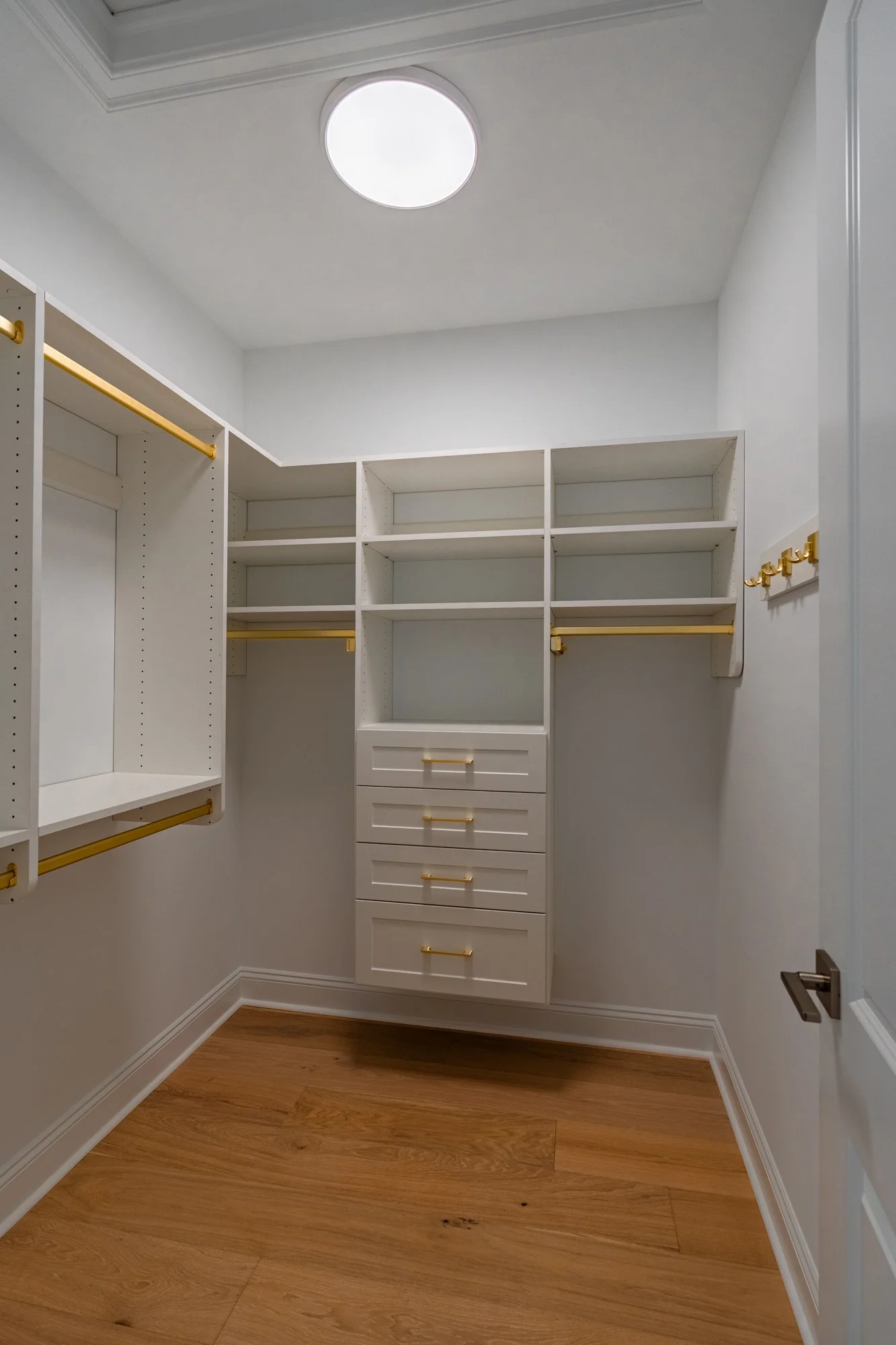 Empty walk-in closet with white shelves and drawers, gold hooks, and wood flooring.