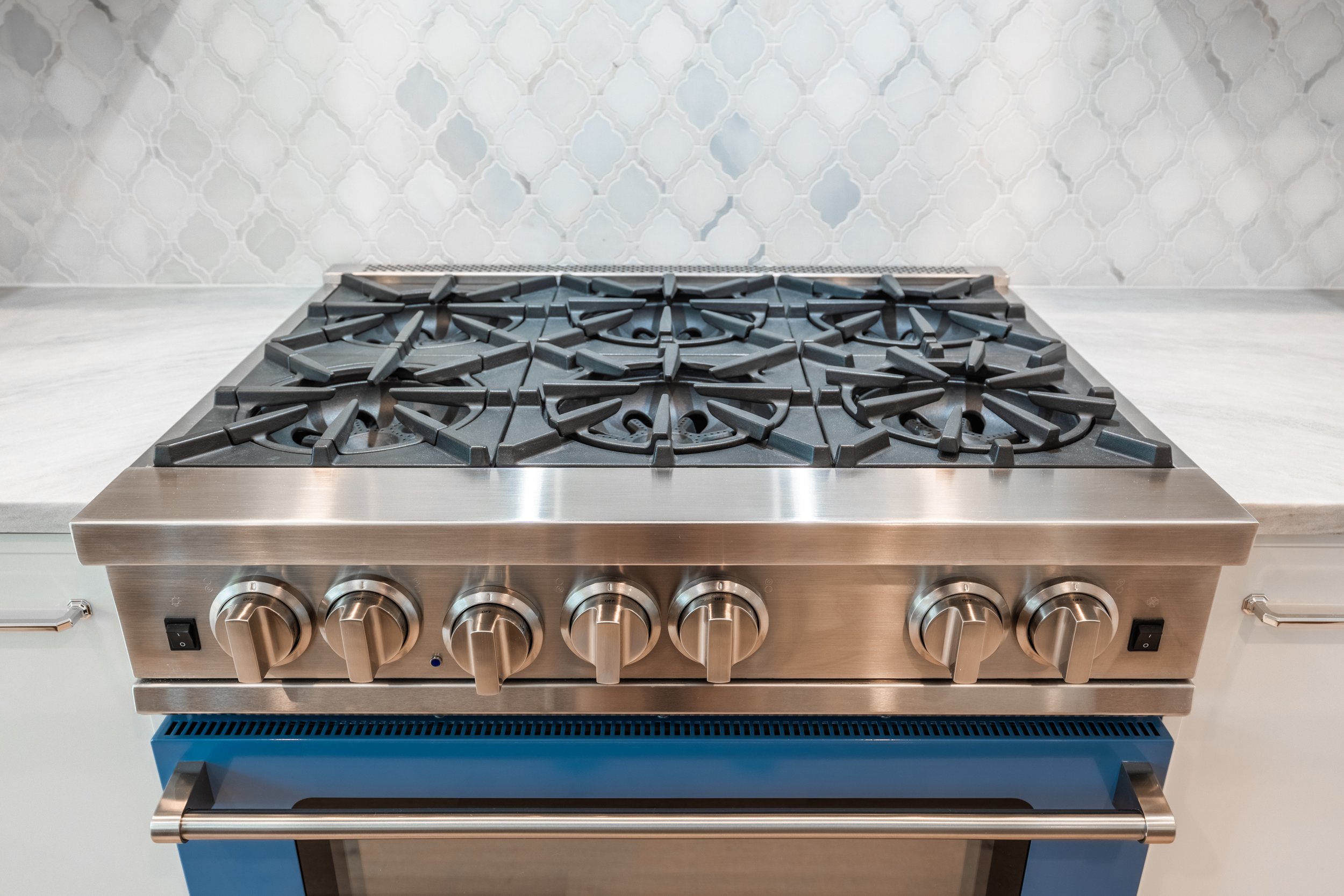 Stainless steel gas stove with six burners and control knobs, set against a white marble countertop and a white decorative tile backsplash.