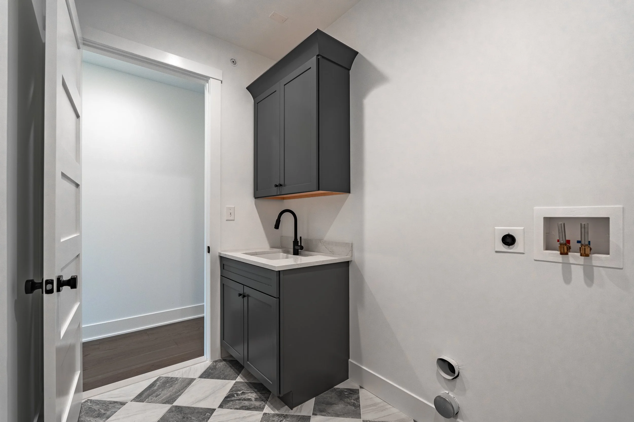 Empty laundry room with dark gray cabinets, white walls, and a black sink faucet. Tile flooring in white, black, and gray diamond pattern, with laundry hookups on the right wall.