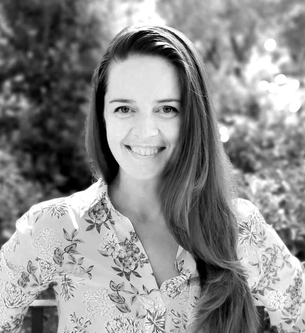 Black and white photo of a smiling woman with long hair, wearing a floral shirt, outdoors with blurred natural background.